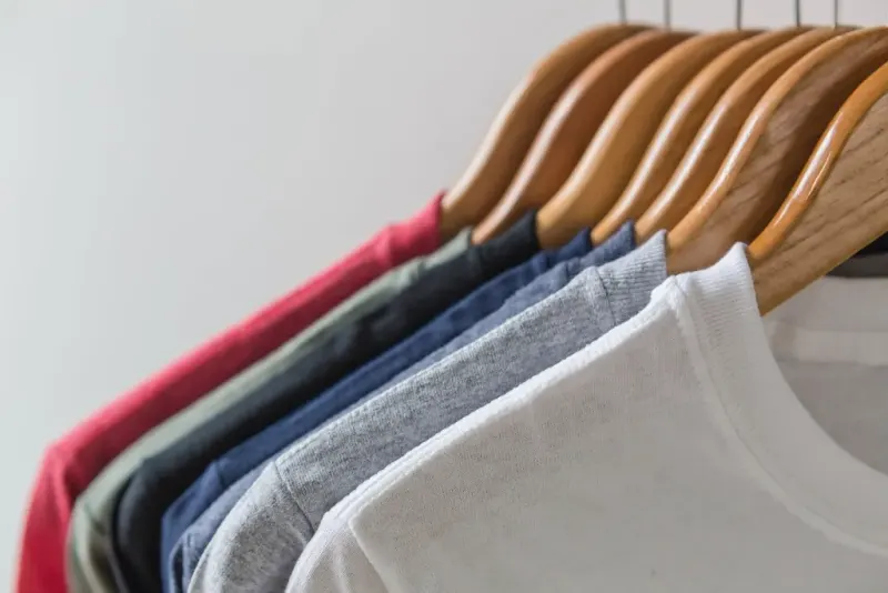 Deretan kaus polos warna putih, abu-abu, biru, hitam, hijau zaitun, dan merah di gantungan kayu, tekstur rajutan dan kerah rib terlihat dari dekat.