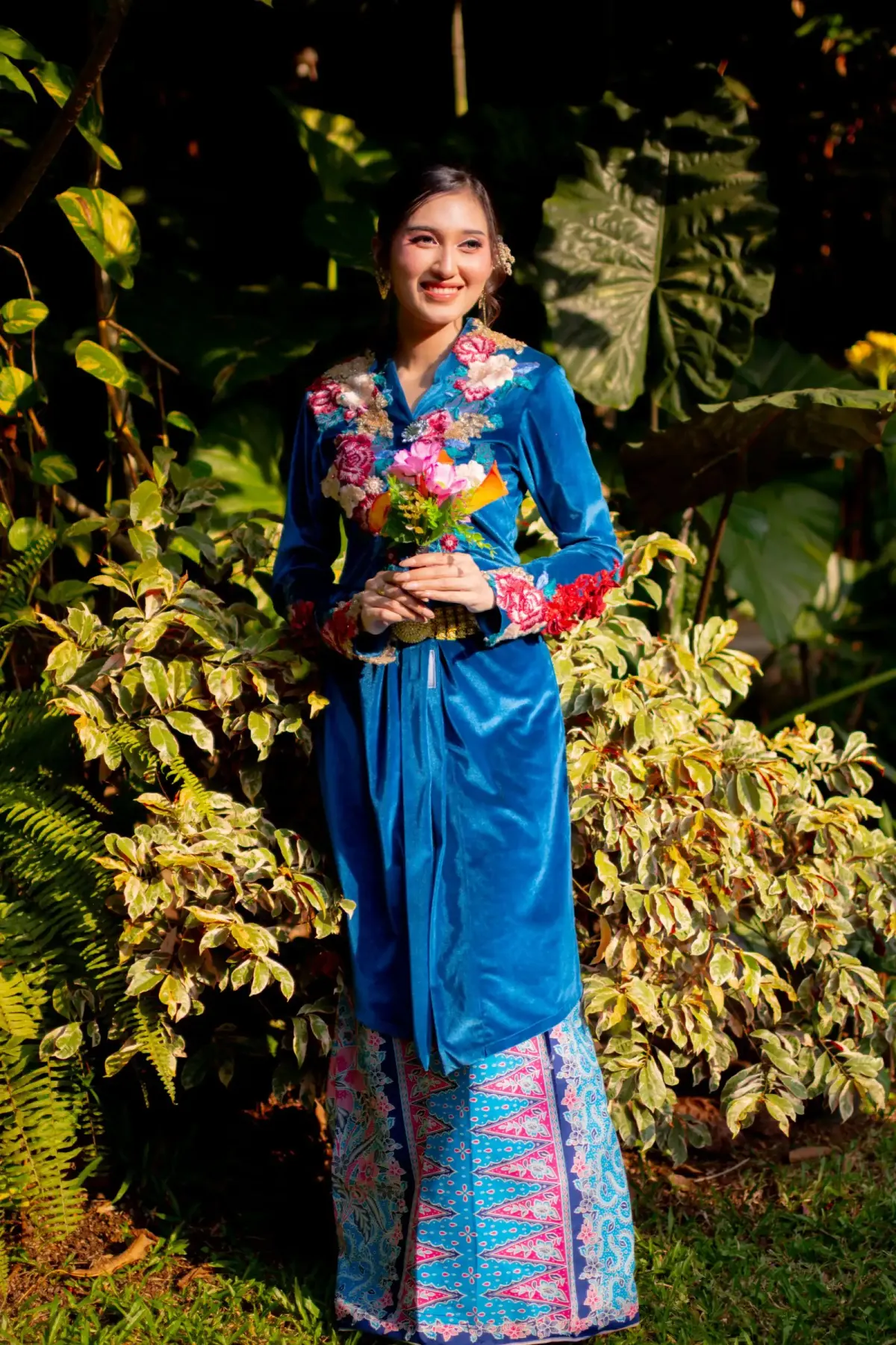 Perempuan berambut disanggul memakai kebaya biru mengilap lengan panjang berbordir bunga di kerah dan manset serta kain bermotif tumpal merah muda dan biru, memegang buket bunga di antara tanaman.