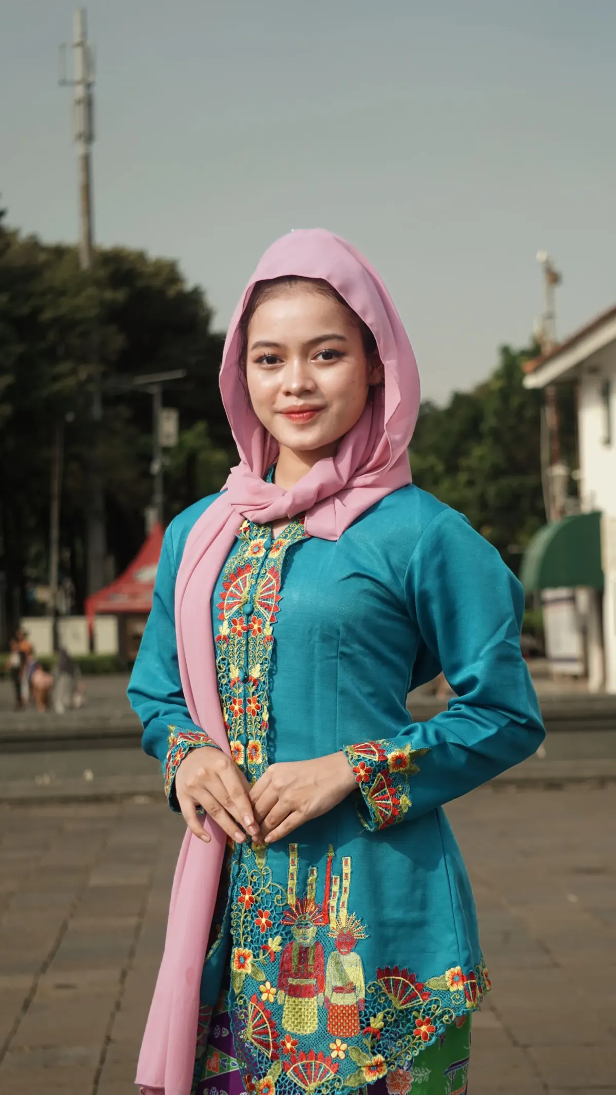 Perempuan berhijab pashmina merah muda memakai kebaya tosca lengan panjang dengan bordir warna-warni di sepanjang bukaan depan dan ujung lengan, berdiri di pelataran kota.