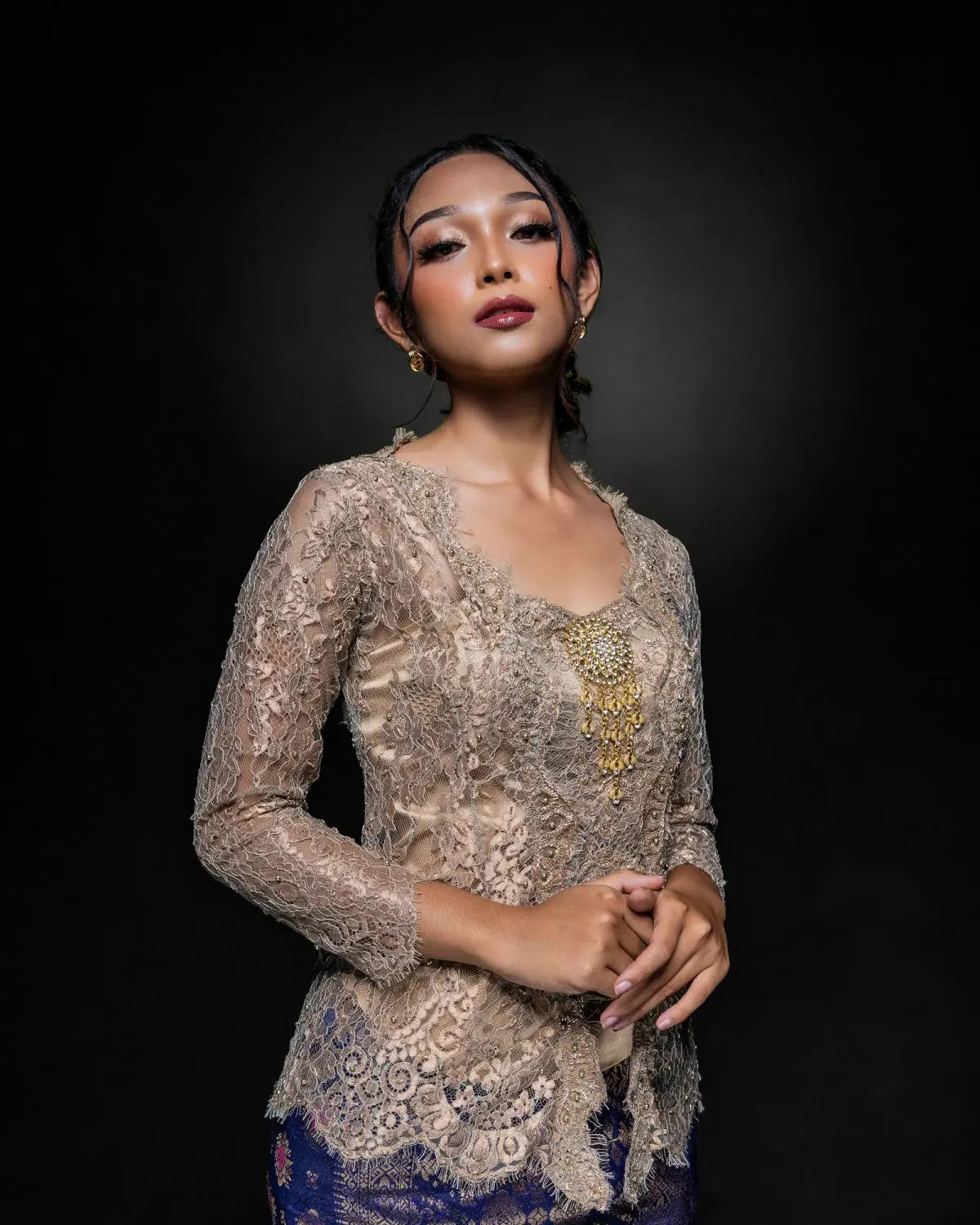 Perempuan bersanggul memakai kebaya brokat beige keemasan lengan tiga perempat berkerah V dengan bros emas di dada, dipadukan kain bermotif biru tua, di depan latar studio gelap.