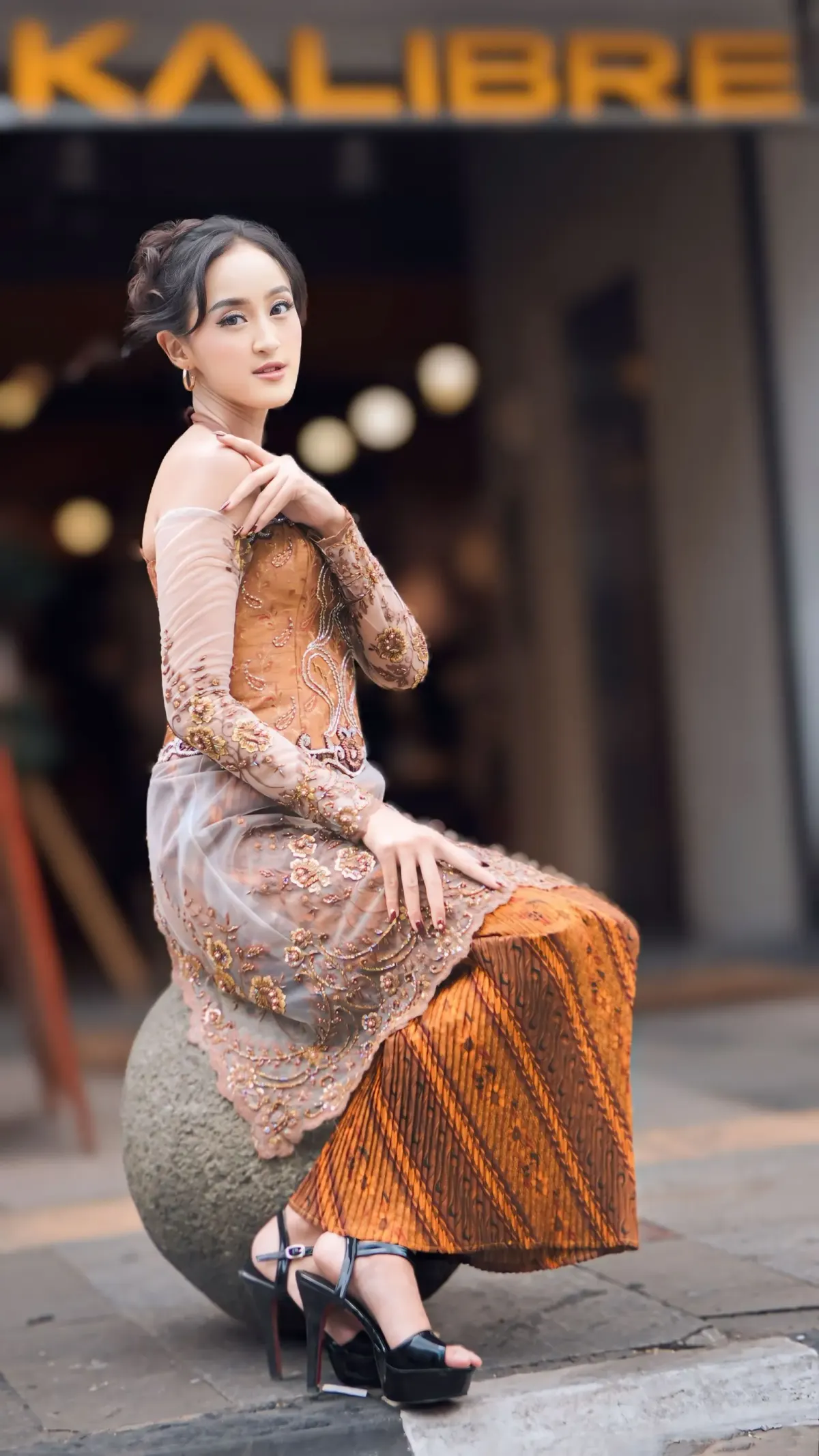 Perempuan bersanggul memakai kebaya brokat tipis warna cokelat susu berpayet dengan bahu terbuka, kain batik cokelat jingga, dan sepatu platform hitam, duduk di bola batu di depan toko.