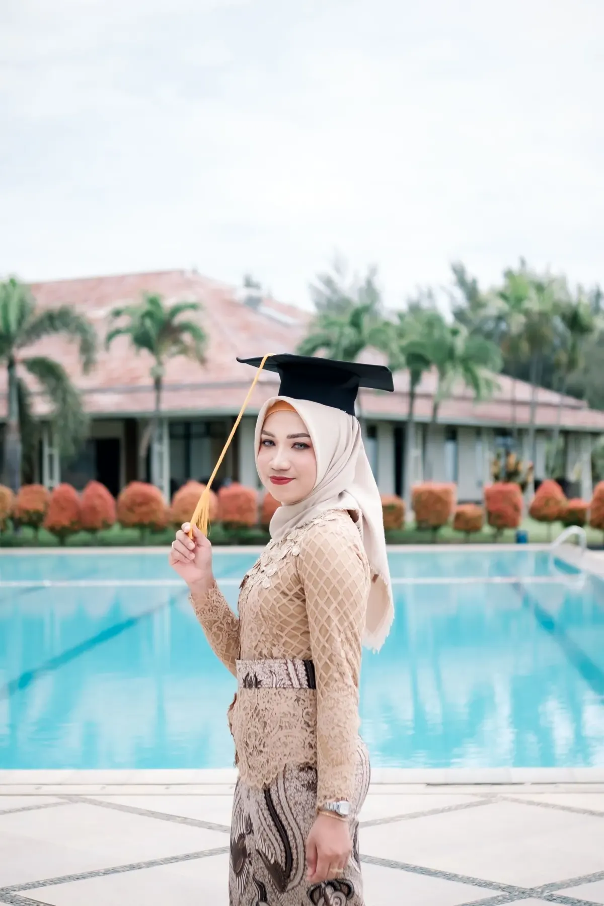 Perempuan berhijab krem memakai kebaya brokat krem lengan panjang dengan ikat pinggang motif, kain batik cokelat, dan topi toga hitam berumbai emas, berpose di tepi kolam renang.