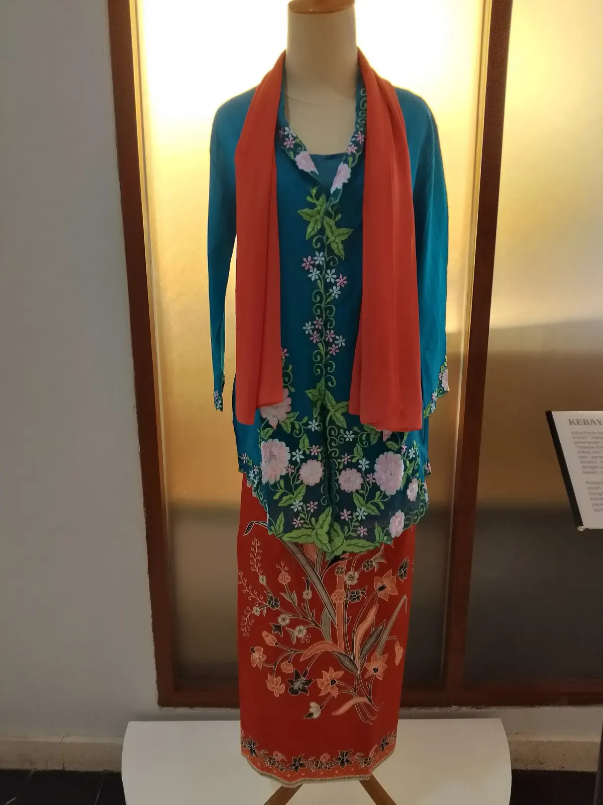 Kebaya encim biru toska berbordir bunga merah muda dan daun hijau di tepi depan, dipajang pada manekin dengan selendang oranye dan kain batik merah bermotif bunga.