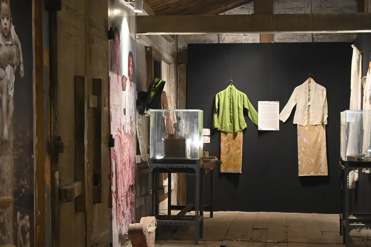 Dua kebaya encim, hijau dan krem, tergantung bersama kain batik di dinding hitam ruang pamer museum berlantai dan berdinding kayu.