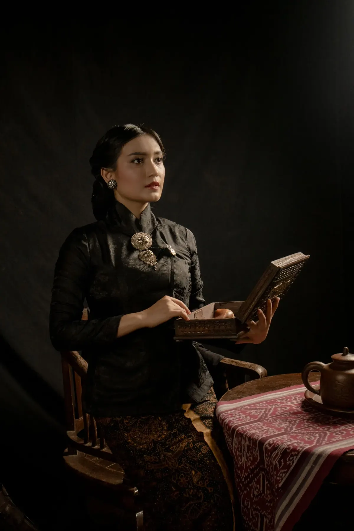 Perempuan duduk memegang kotak kayu berukir, memakai kebaya hitam bertekstur dengan kerah berdiri dan bros logam besar di dada, dipadukan kain batik cokelat tua, berlatar gelap.