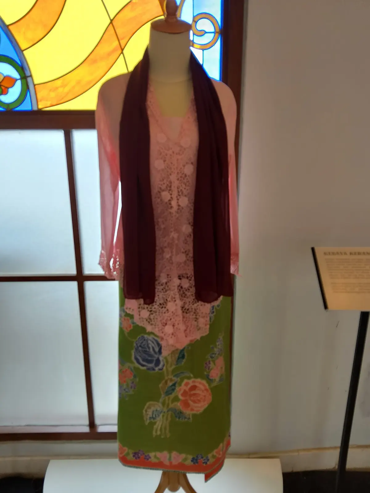 Kebaya kerancang merah muda dengan bordir berlubang bermotif bunga di bagian depan, dipajang pada manekin dengan selendang merah tua dan kain batik hijau bermotif mawar.