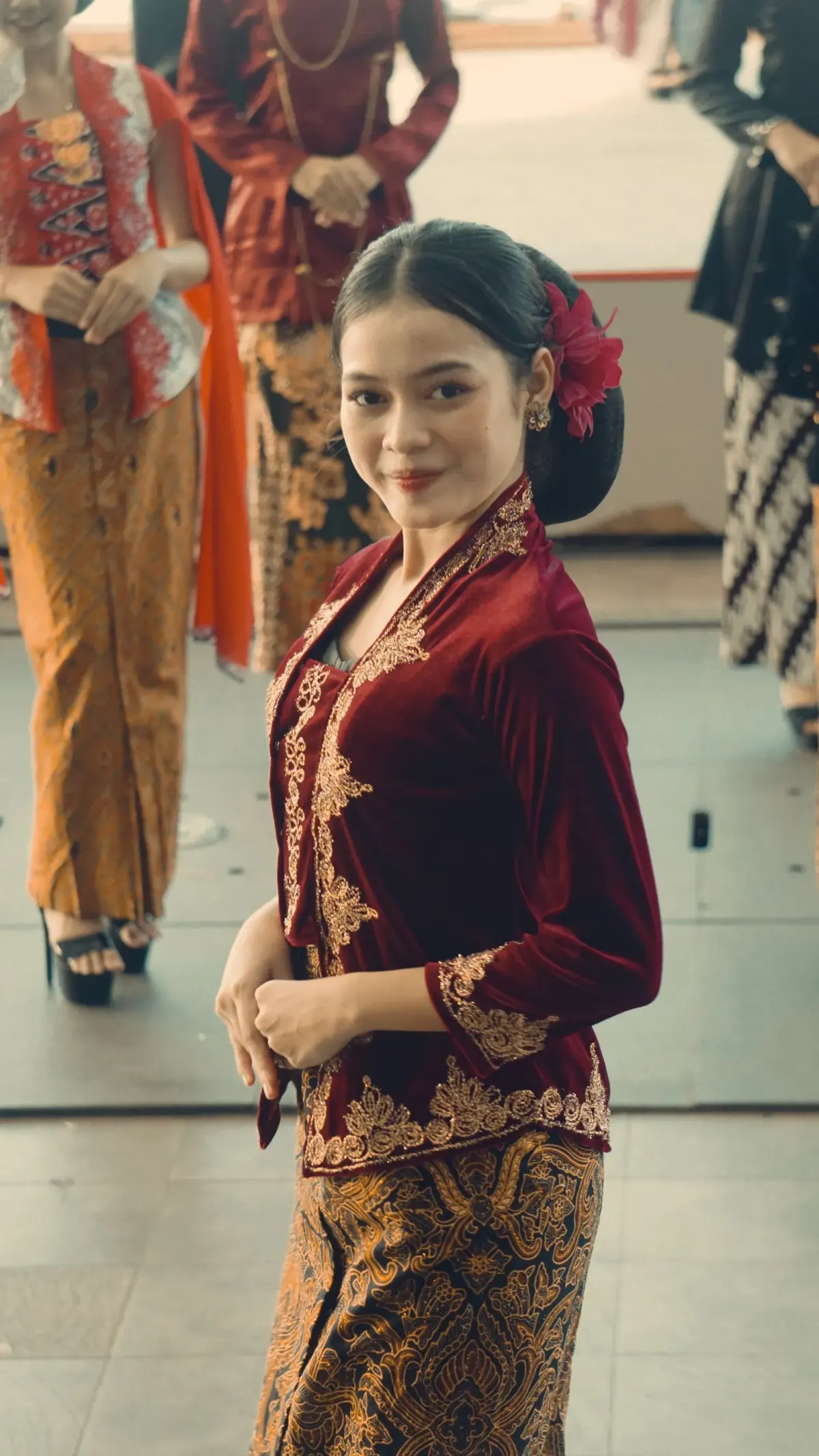 Perempuan bersanggul dengan bunga merah memakai kebaya merah marun bertekstur lembut berhias bordir emas di tepi depan dan ujung lengan, dipadukan kain batik cokelat keemasan.