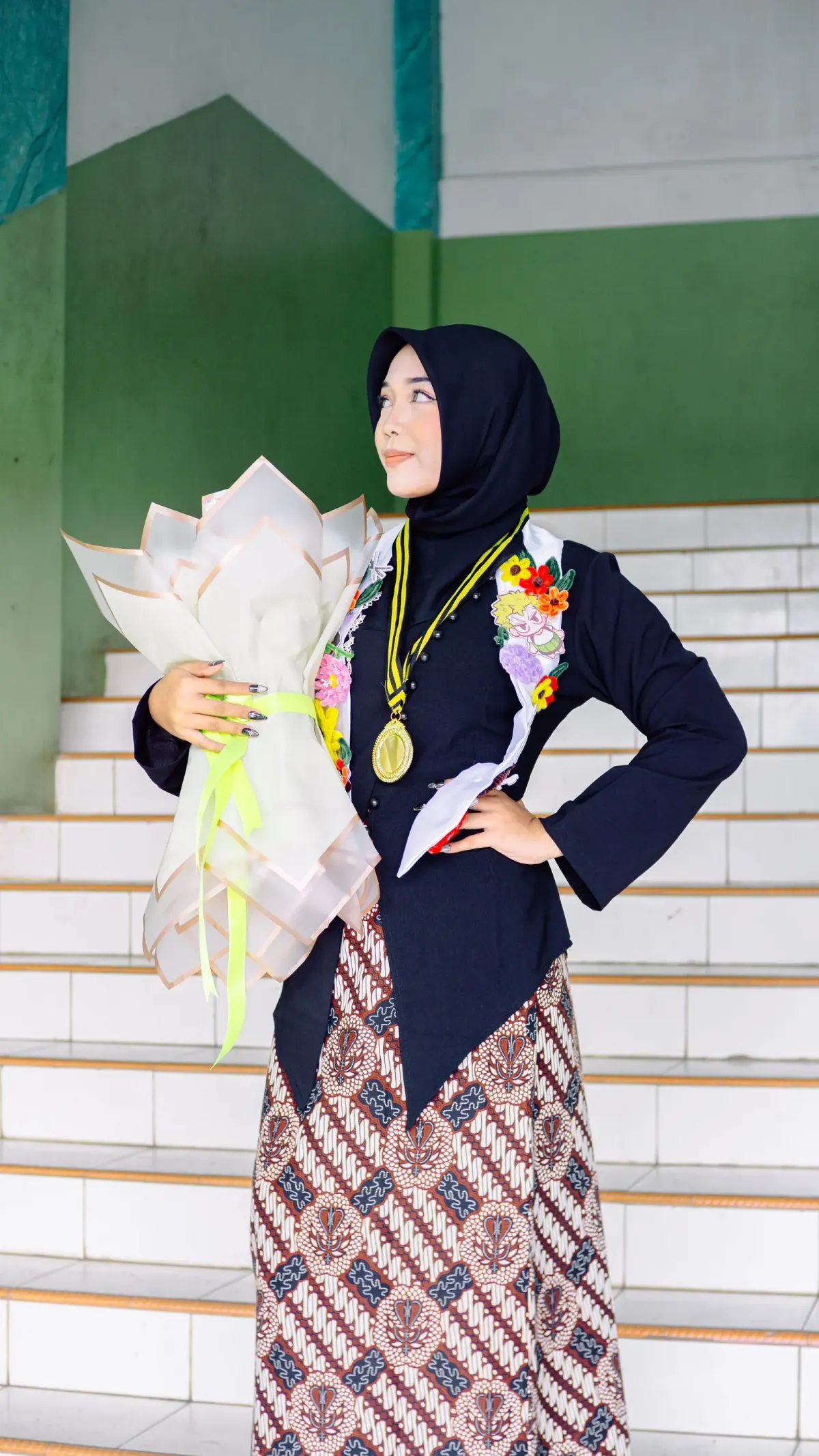 Perempuan berhijab navy memakai kebaya navy lengan panjang dengan kerah putih berbordir bunga, medali wisuda, dan rok batik krem bermotif cokelat, memeluk buket bunga kertas di tangga.