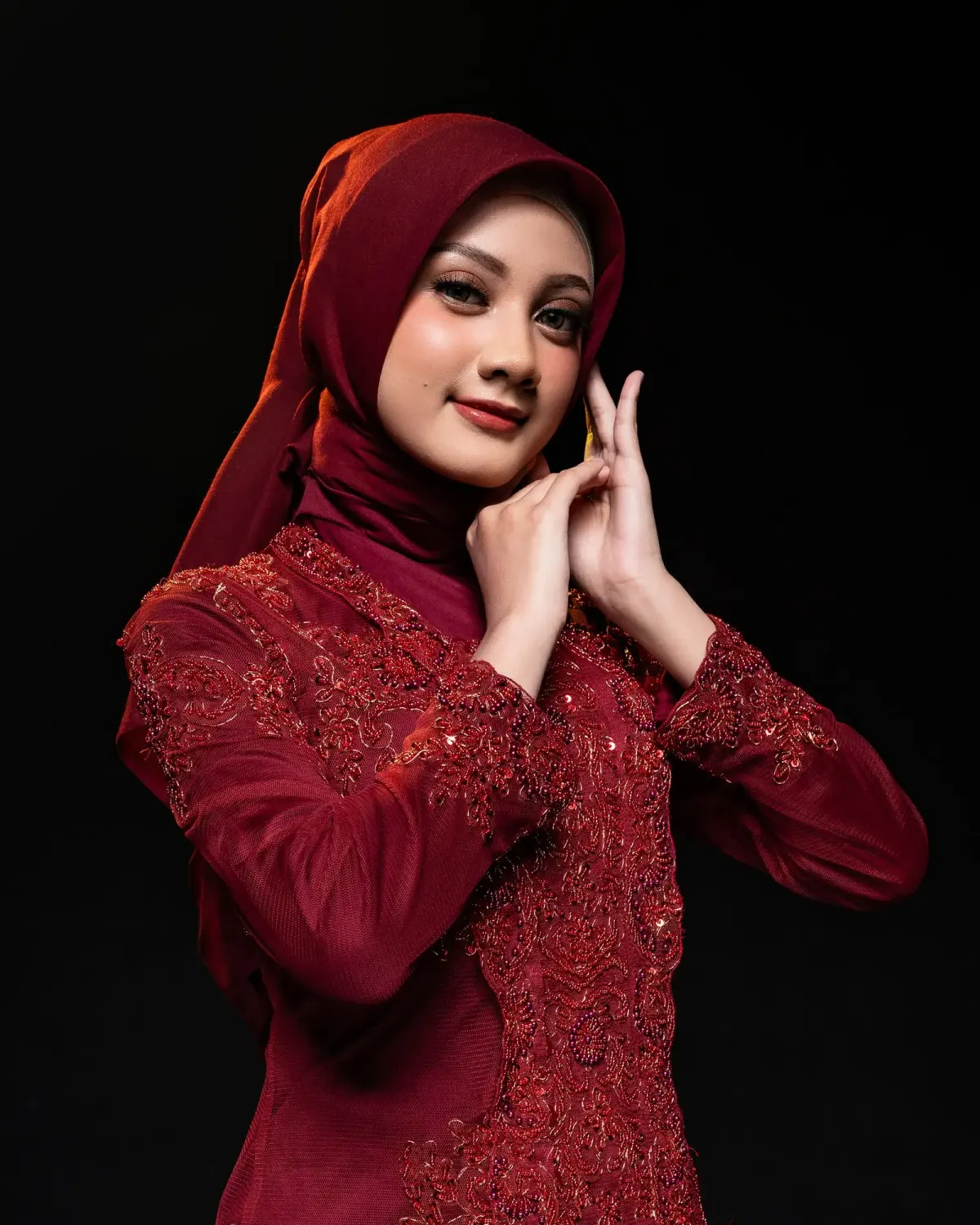 Perempuan berhijab maroon memakai kebaya lengan panjang maroon berhias renda dan payet senada di dada dan ujung lengan, berpose di depan latar studio hitam.