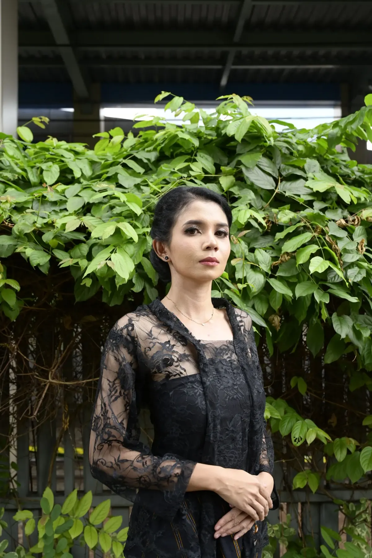 Perempuan berkebaya renda hitam lengan panjang yang dipakai terbuka di atas dalaman hitam, tangan dilipat di depan badan, berlatar tanaman merambat hijau.