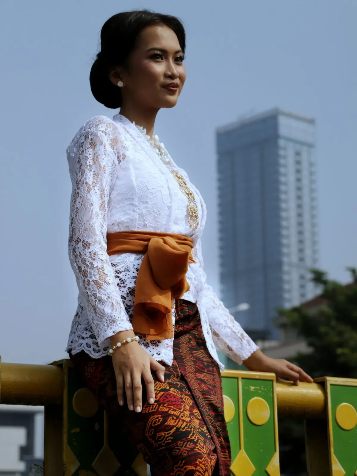 Perempuan berkebaya renda putih lengan panjang dengan hiasan emas di bagian depan, selendang oranye diikat di pinggang, dan kain lilit bermotif merah cokelat, berdiri di depan gedung tinggi.
