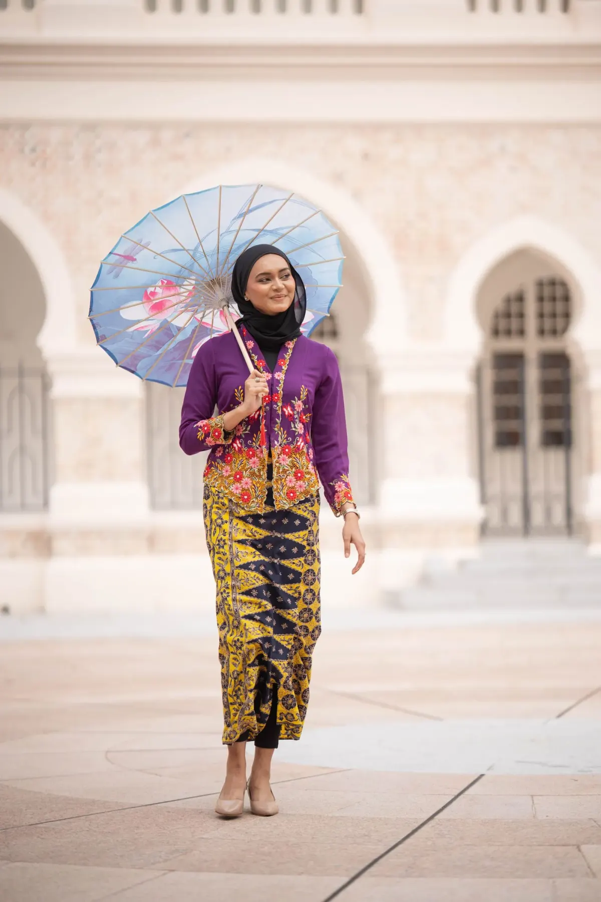 Perempuan berhijab hitam memakai kebaya ungu berbordir bunga merah, kain batik kuning bermotif ungu, dan sepatu hak krem, memegang payung kertas bermotif di depan bangunan berlengkung.