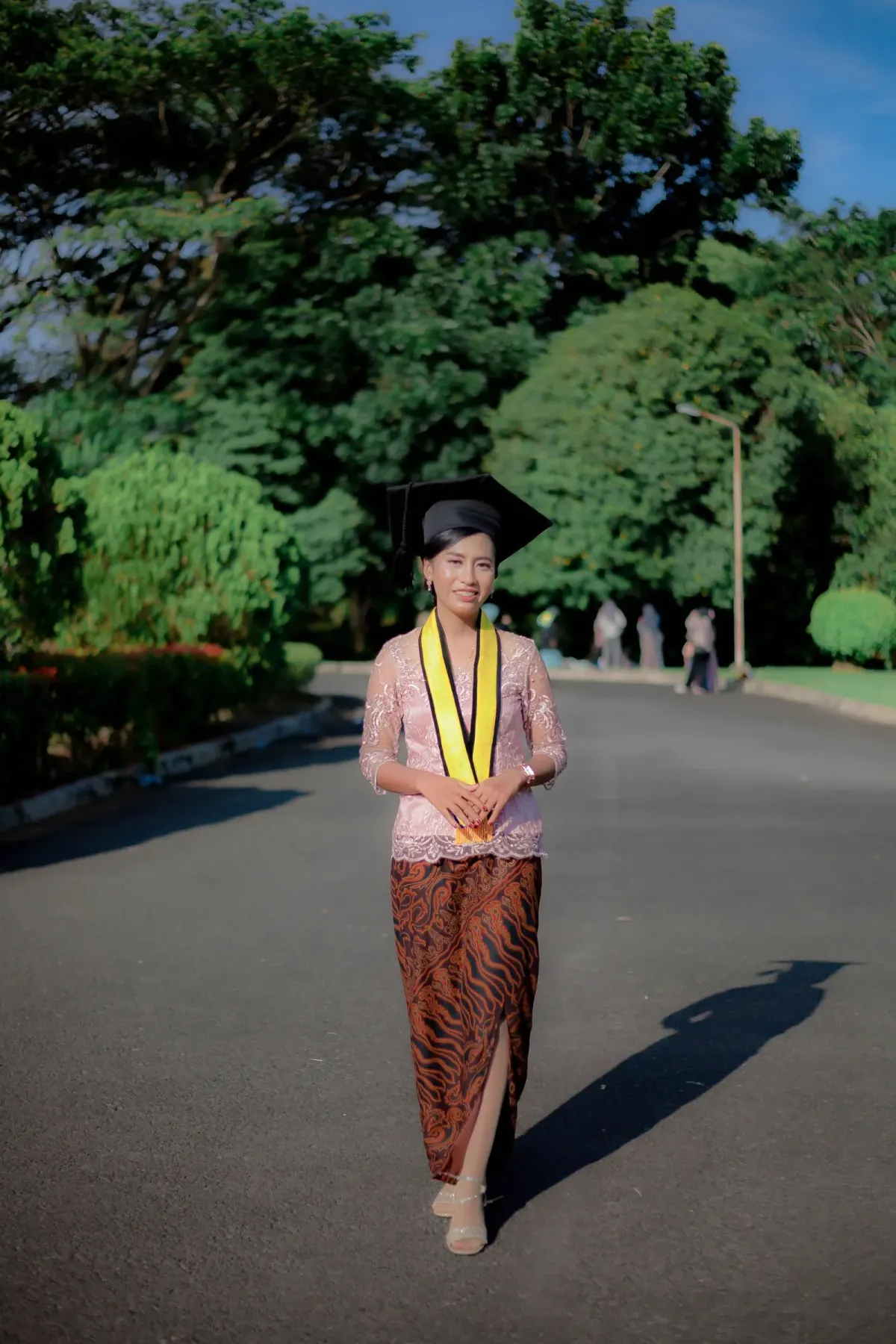 Wisudawati bertopi toga hitam dan selempang kuning memakai kebaya brokat merah muda lengan tiga perempat dengan bawahan kain batik cokelat berbelahan samping dan sandal hak keemasan, berjalan di jalan kampus berpohon rindang.