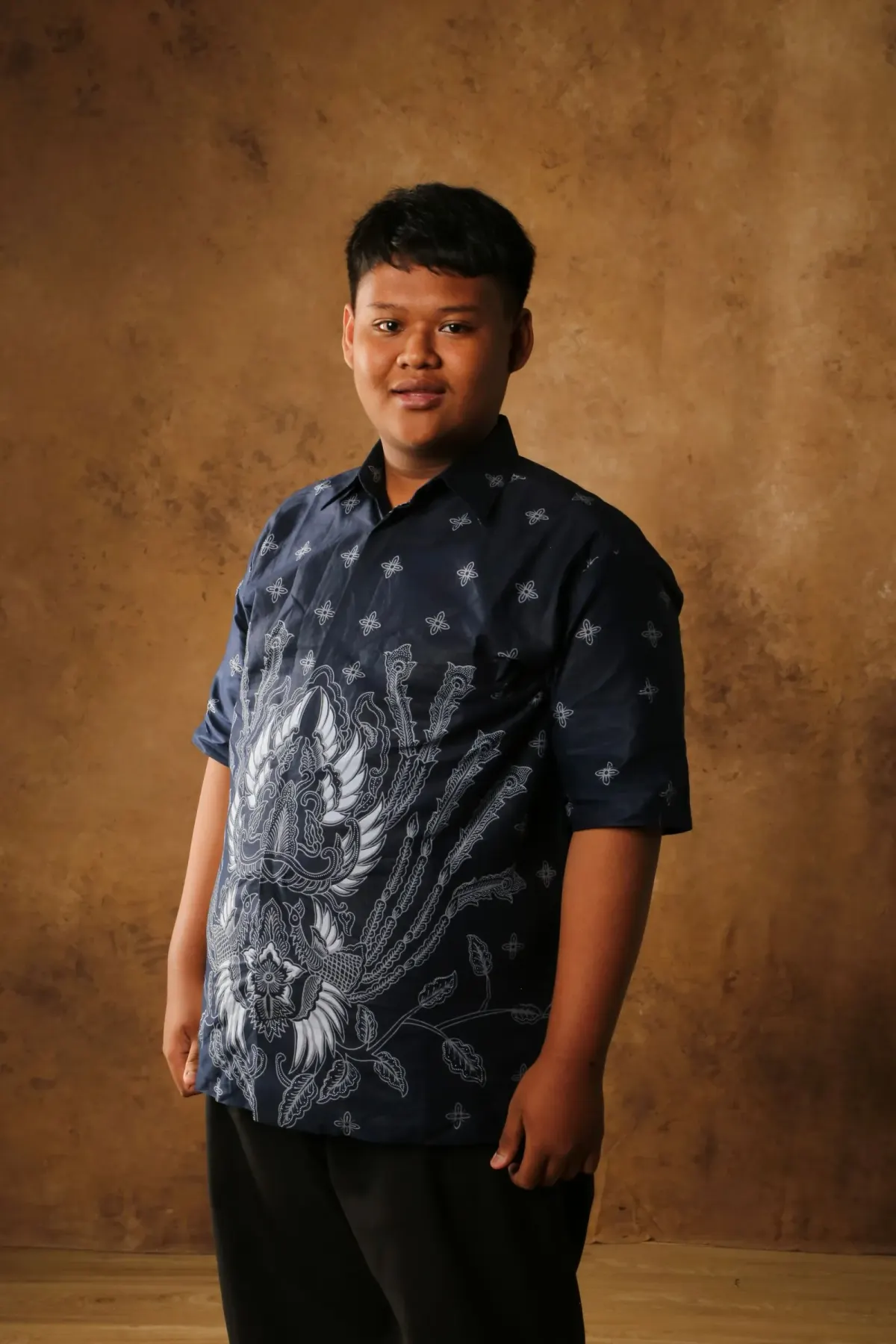 Pria muda memakai kemeja batik lengan pendek berwarna navy dengan motif burung dan bunga putih besar, celana hitam, berpose di depan latar studio cokelat.