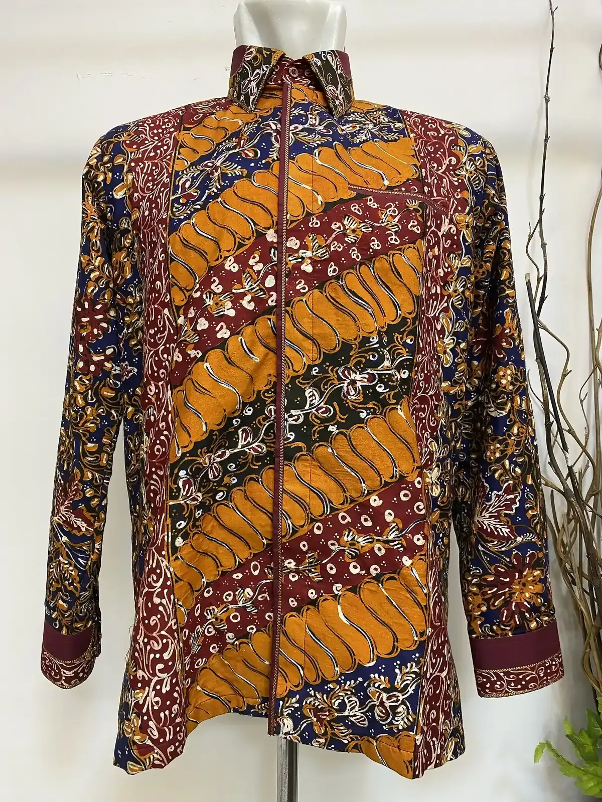 Kemeja batik lengan panjang pada manekin, motif diagonal oranye, merah marun, biru tua, dan hijau gelap, dengan kerah dan manset berlis merah marun.