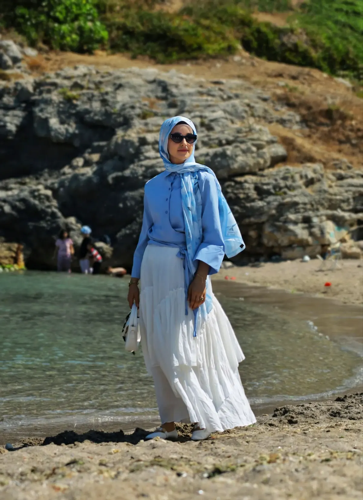 Perempuan berhijab biru motif dan berkacamata hitam memakai kemeja biru diikat di depan, rok panjang putih bertumpuk, sepatu putih, dan tas kecil putih, berdiri di pantai berkarang.
