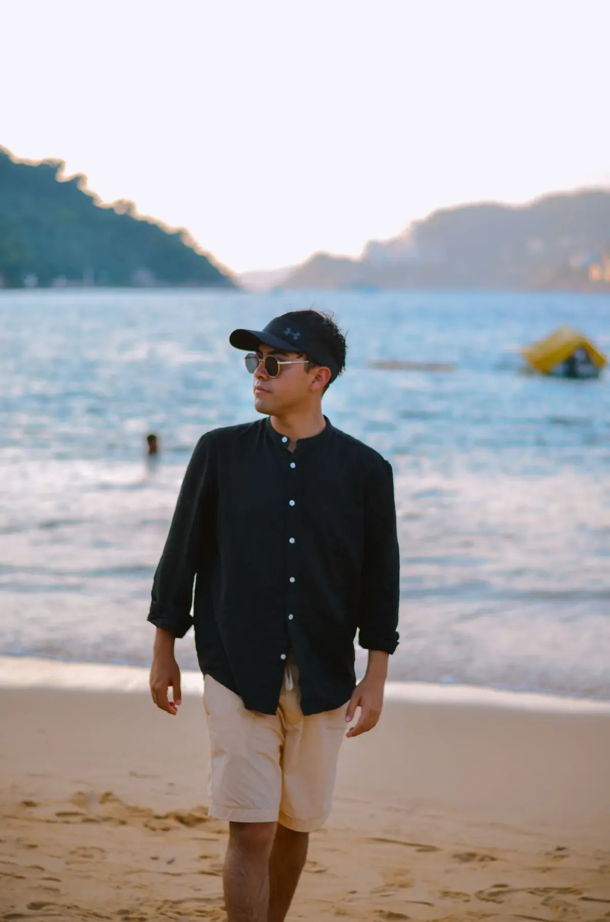 Pria bertopi hitam dan berkacamata hitam memakai kemeja hitam lengan panjang berkerah pendek yang digulung dan celana pendek krem, berjalan di pantai berpasir.