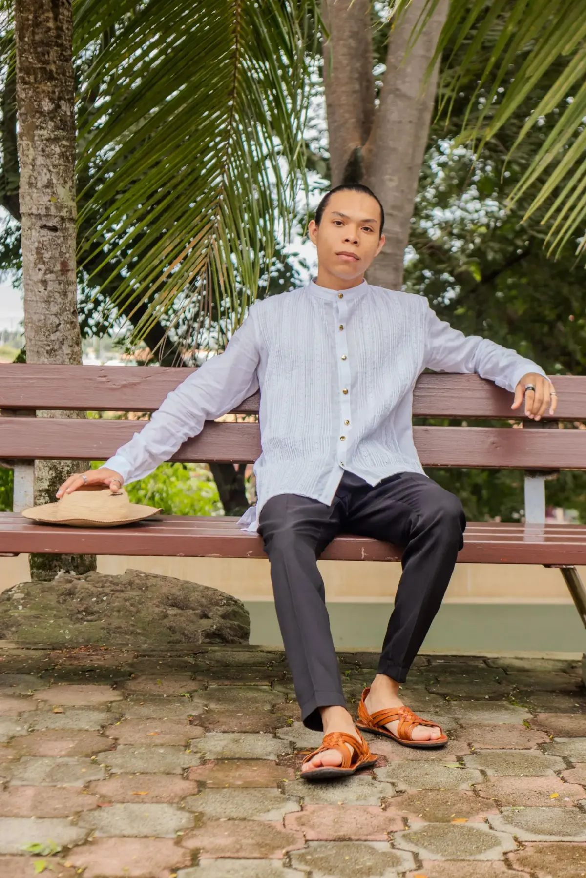 Pria memakai kemeja lengan panjang abu-abu muda berkerah pendek, celana panjang abu-abu tua, dan sandal kulit cokelat, duduk di bangku taman di bawah pohon kelapa dengan topi jerami di sampingnya.