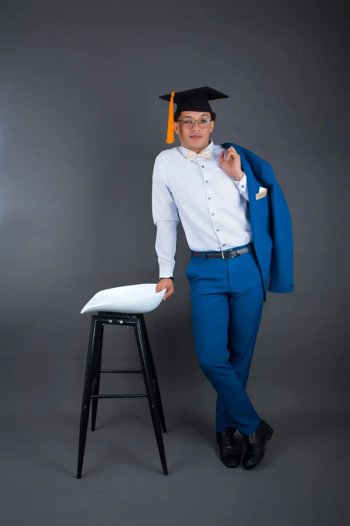 Pria berkacamata dan bertopi toga memakai kemeja putih, dasi kupu-kupu krem, celana biru, sabuk hitam, dan sepatu hitam, menyampirkan jas biru di bahu, berpose di studio abu-abu.