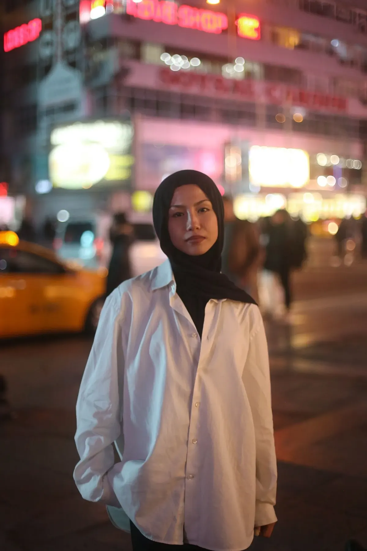 Perempuan berhijab hitam memakai kemeja putih oversized lengan panjang, berdiri di trotoar kota pada malam hari dengan lampu toko dan taksi kuning di latar belakang; lokasinya tidak disebut sumber dan tampaknya bukan Indonesia.