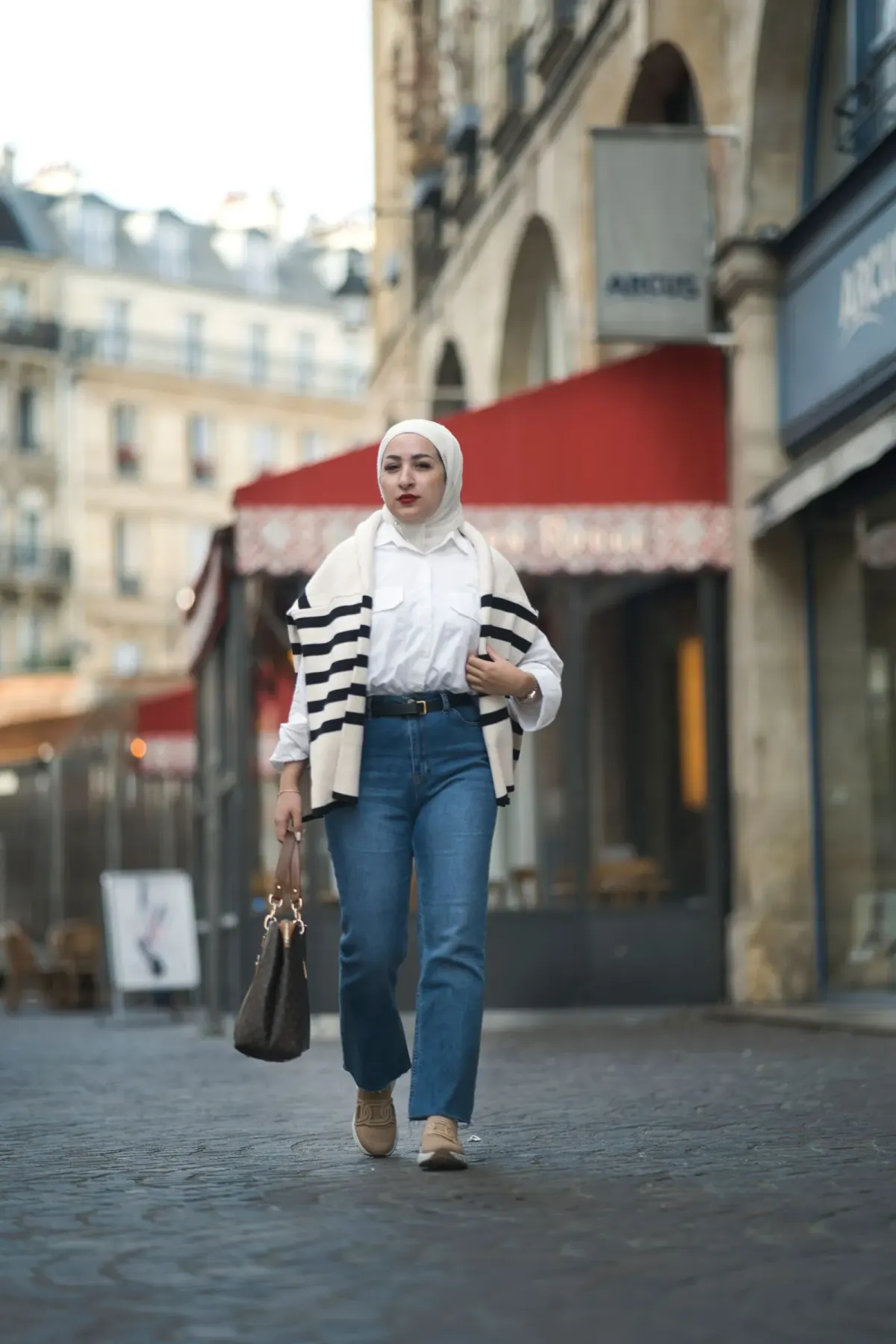 Perempuan berhijab putih memakai kemeja putih, kardigan krem bergaris hitam disampirkan di bahu, jins biru lurus bersabuk hitam, sepatu slip-on beige, dan tas tangan cokelat tua bermotif, berjalan di jalan kota Eropa.