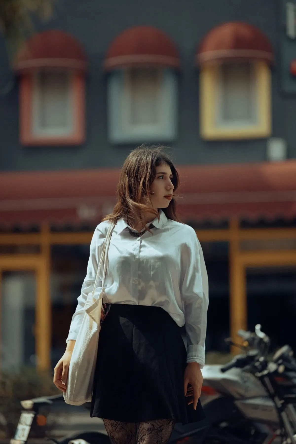 Perempuan berambut panjang memakai atasan putih berkerah lengan panjang yang dimasukkan ke rok pendek hitam, membawa tas tote kanvas krem di jalan kota.