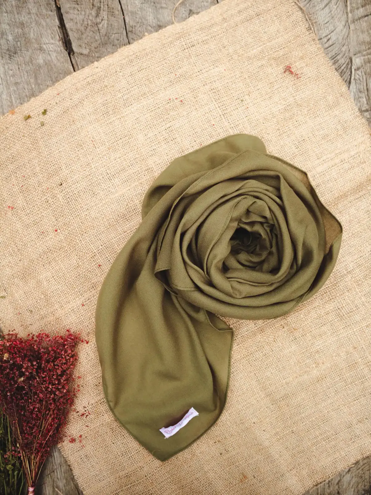 Kerudung panjang polos warna hijau zaitun digulung melingkar di atas kain goni dan papan kayu, dengan seikat bunga kering merah di sudut.
