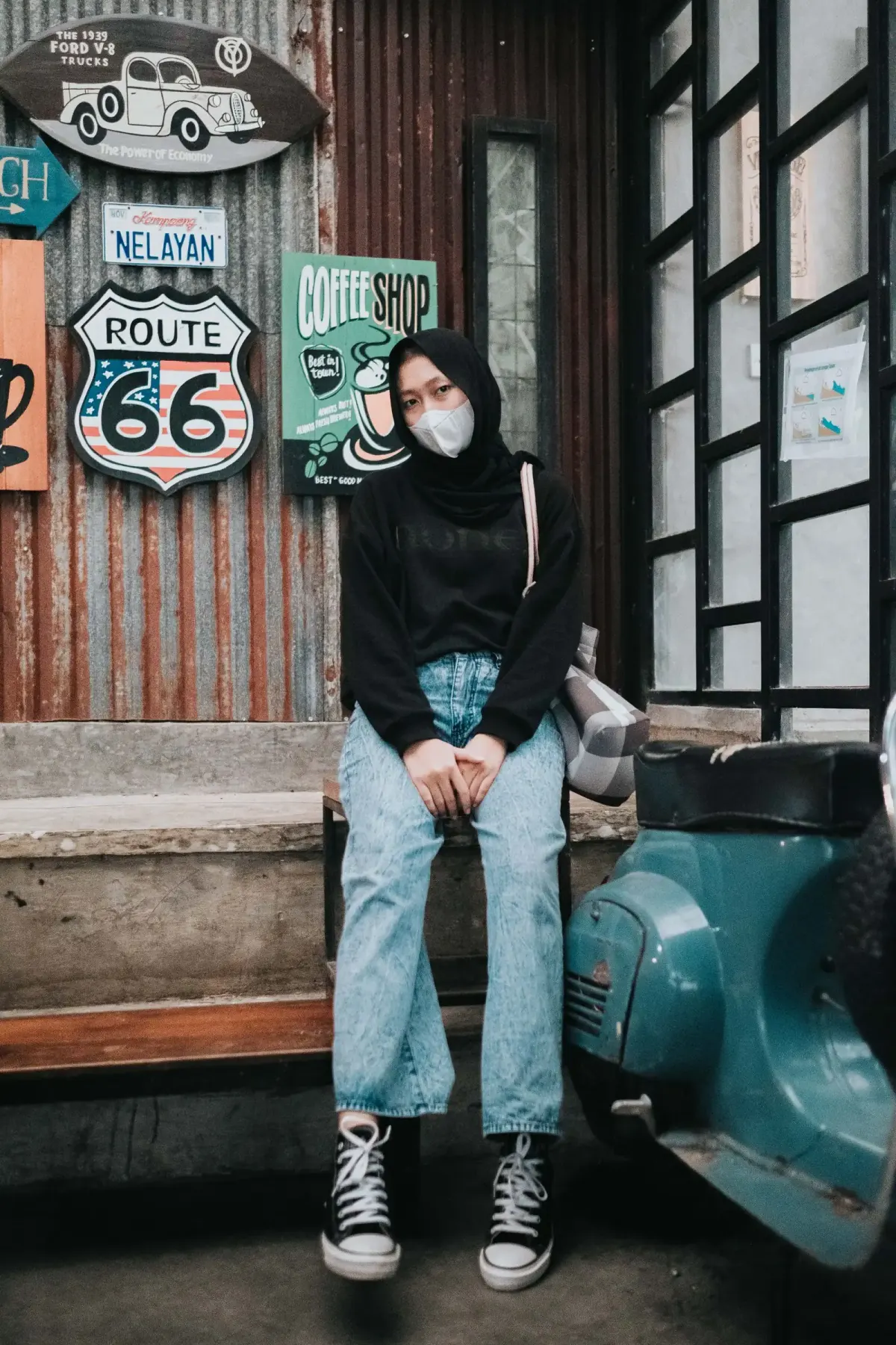 Perempuan berhijab hitam dan bermasker putih duduk di bangku kayu, memakai sweter hitam, celana jins biru muda berpotongan lurus longgar, sepatu kets high-top hitam bertali putih, dan tas bahu bermotif kotak abu-abu putih.