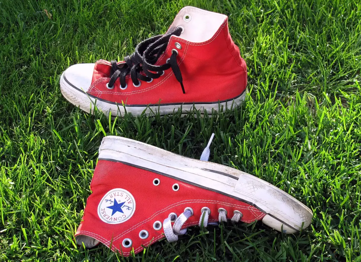 Sepasang sepatu kets kanvas high-top merah dengan sol karet putih, ujung sepatu karet, dan tali hitam serta putih, tergeletak di atas rumput hijau.