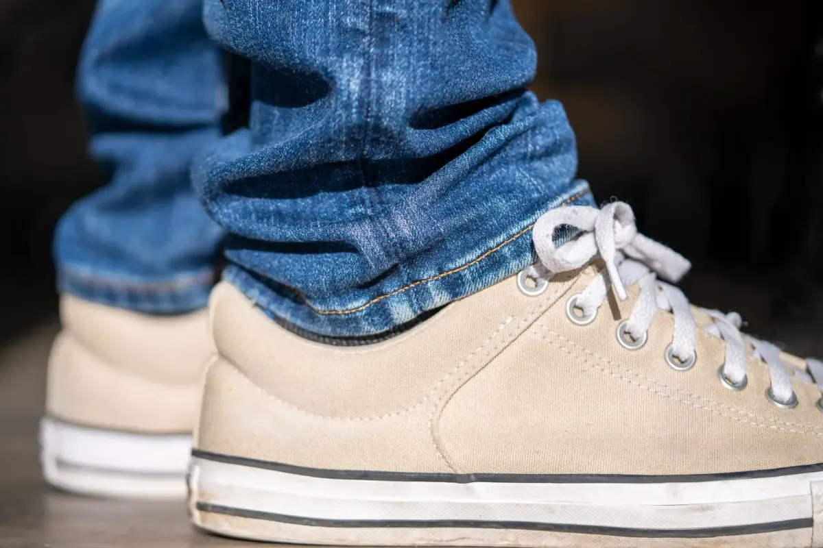 Close-up sepatu kets kanvas krem model low-top dengan tali putih dan sol karet putih bergaris hitam, dipakai dengan celana jins biru yang ujungnya tergulung.