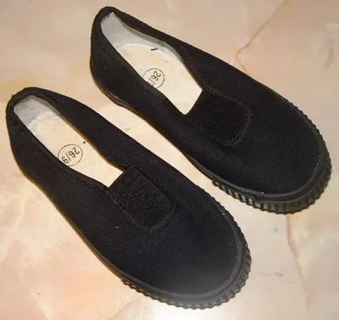 Sepasang sepatu kanvas hitam anak tanpa tali dengan panel karet elastis di sisi lidah sepatu dan sol karet hitam bergerigi, diletakkan di atas lantai marmer krem.