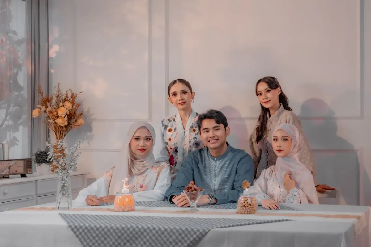 Pria berbaju koko abu-abu kebiruan berkerah berdiri dengan bordir di bagian depan duduk di meja makan bersama empat perempuan berbusana pastel, dengan toples kue dan kurma di atas meja.