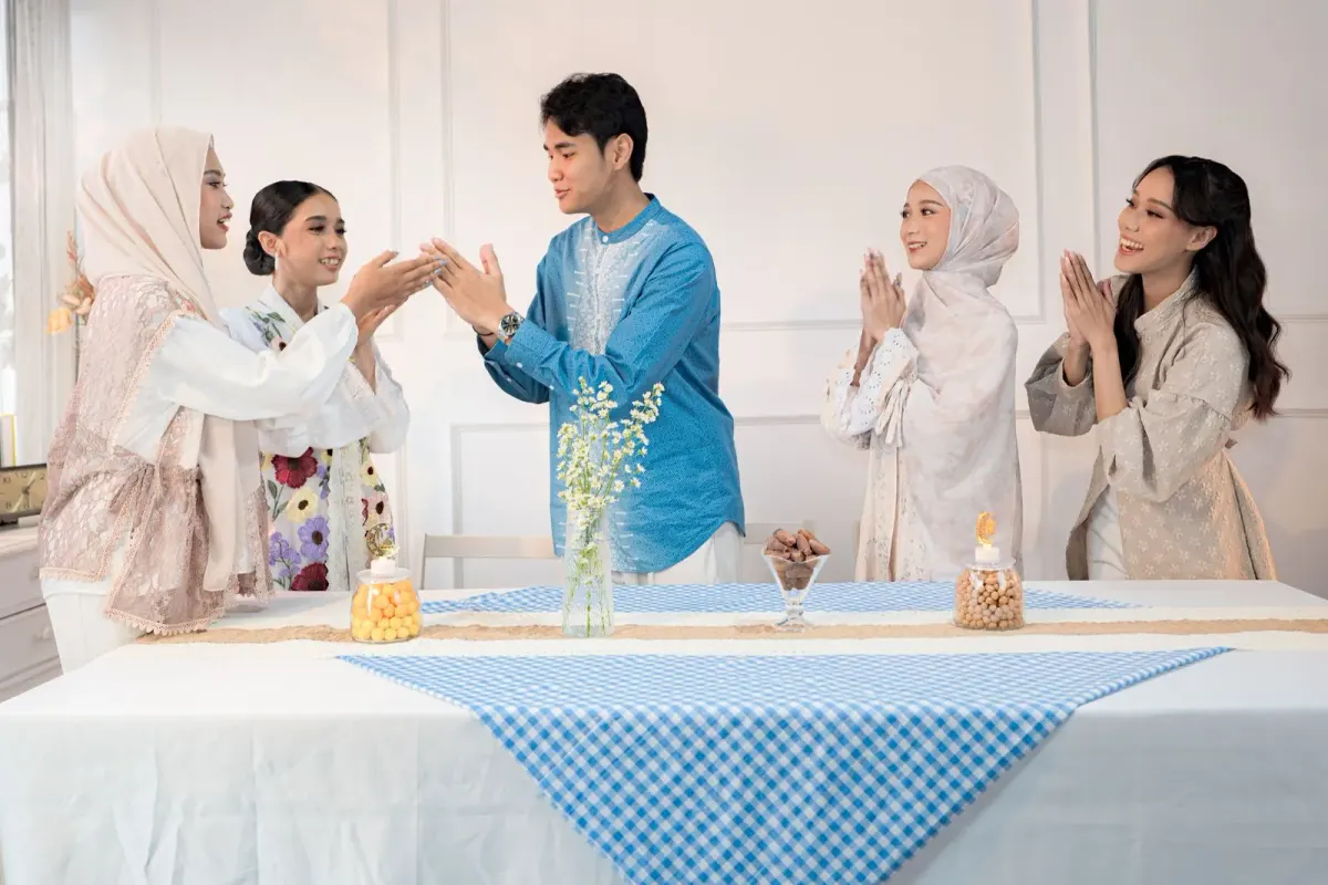 Pria berbaju koko biru lengan panjang dengan bordir putih di bagian depan dan celana putih bersalaman dengan perempuan berhijab di dekat meja bertaplak kotak-kotak biru.