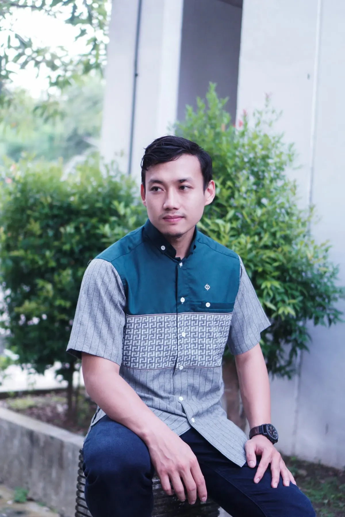 Pria memakai kemeja lengan pendek model koko modern dengan bagian bahu toska dan badan abu-abu bermotif geometris, celana navy, dan jam tangan hitam, duduk di taman, Jawa Timur.