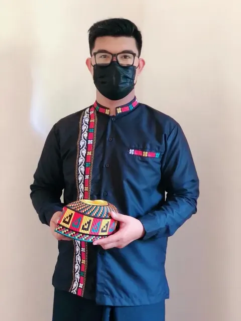 Pria bermasker hitam memakai baju koko navy berkerah berdiri dengan pita motif kerawang Gayo di kerah, sepanjang kancing, dan saku, sambil memegang kopiah bermotif kerawang warna-warni.