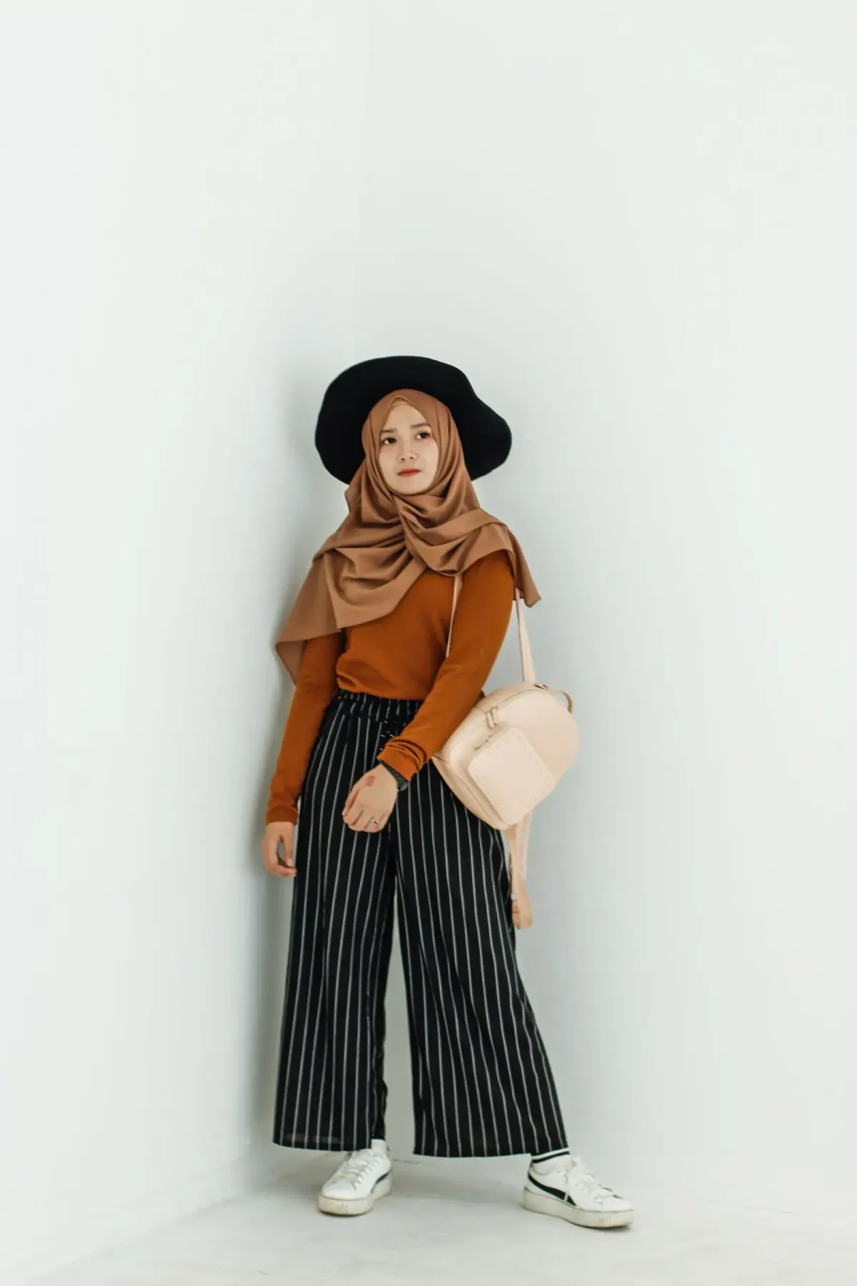 Perempuan berhijab cokelat dan topi hitam lebar memakai atasan lengan panjang cokelat kemerahan, celana kulot bergaris hitam putih sepanjang mata kaki, sepatu kets putih, dan ransel krem, berdiri di sudut ruangan putih.