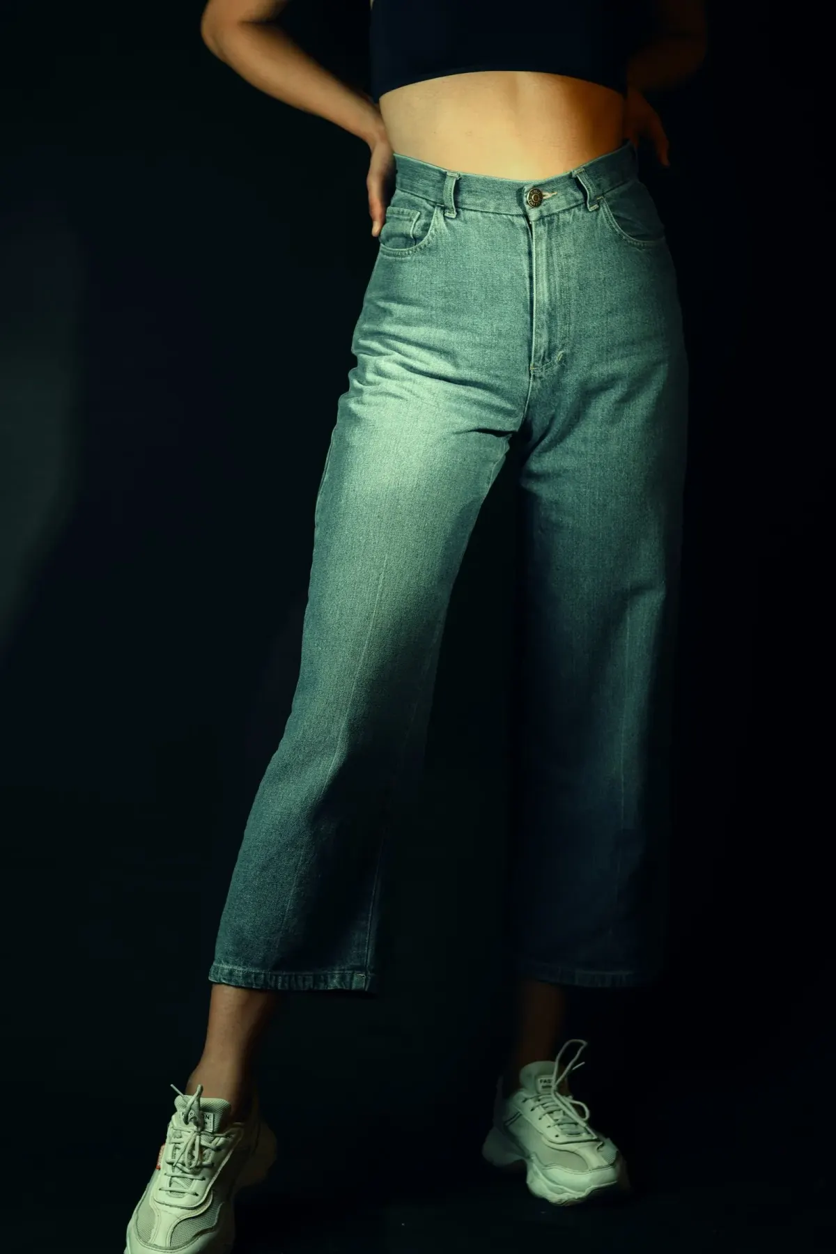 Tampilan pinggang ke bawah seseorang memakai celana jeans biru muda berpinggang tinggi dengan kaki lebar yang berhenti di atas mata kaki, dipadukan sepatu kets hijau muda, berlatar gelap.