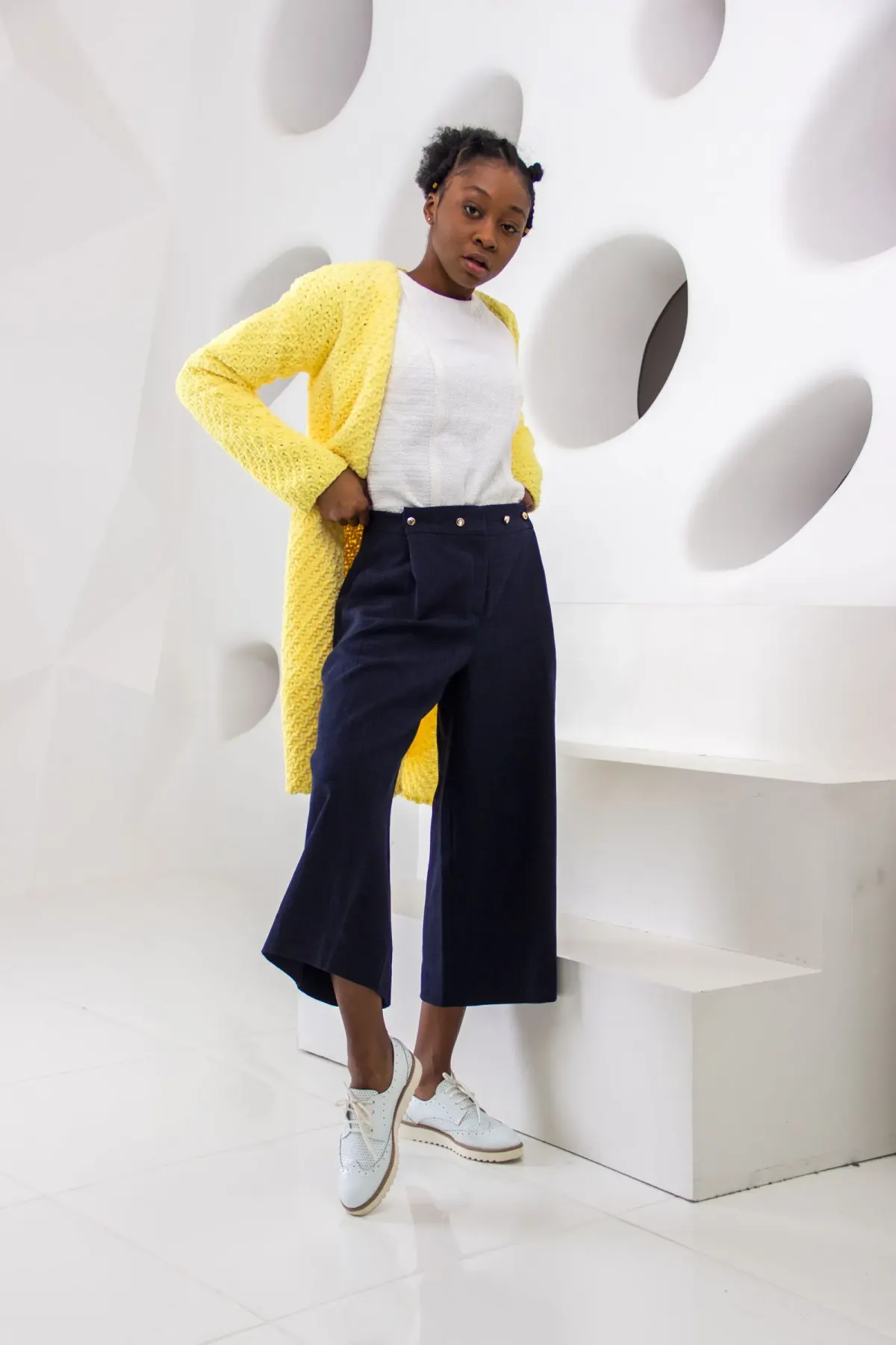 Perempuan memakai celana kulot navy berpinggang tinggi dengan deretan kancing di pinggang, atasan putih, cardigan rajut kuning panjang, dan sepatu oxford putih bersol tebal, berpose di studio putih.