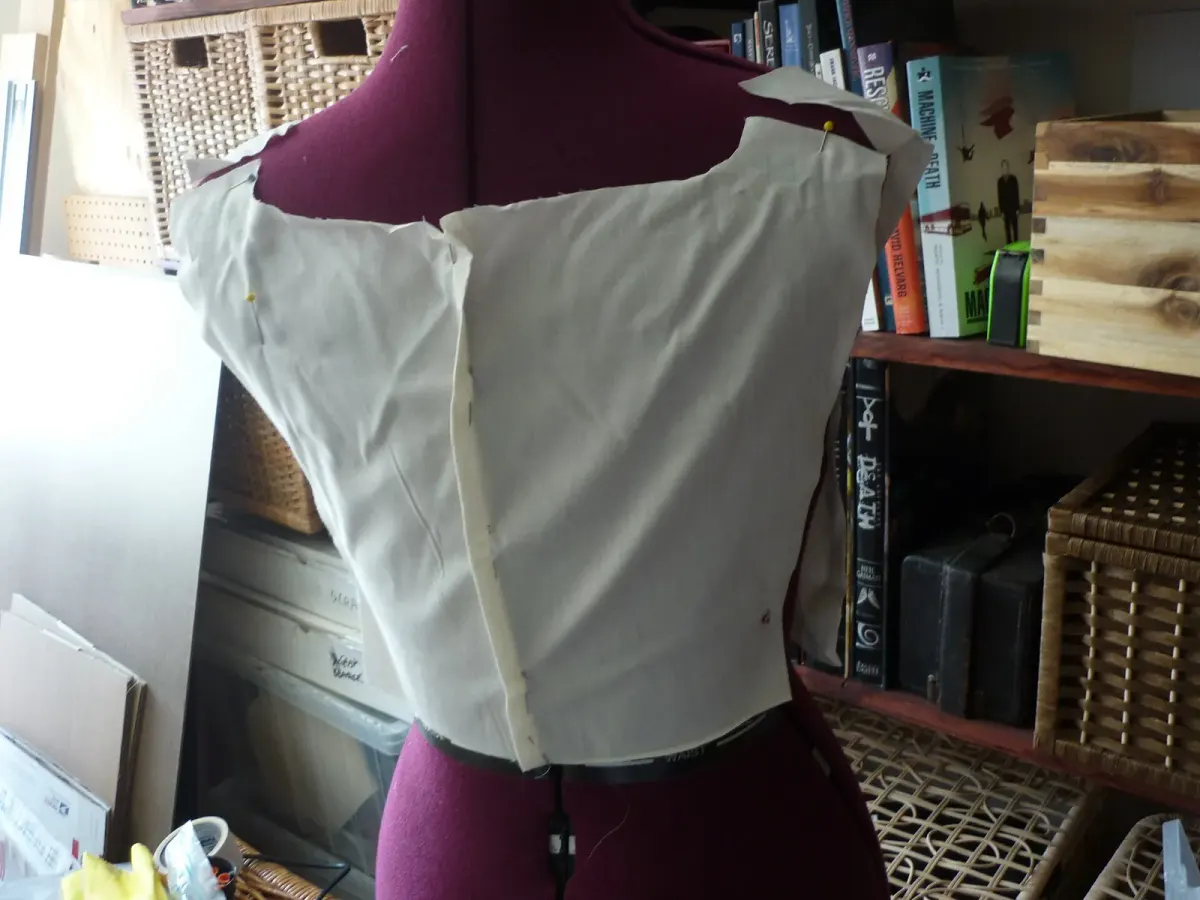 Atasan pola uji dari kain muslin putih gading disematkan dengan jarum pentul pada manekin jahit berwarna merah marun, rak buku dan keranjang anyaman di latar.
