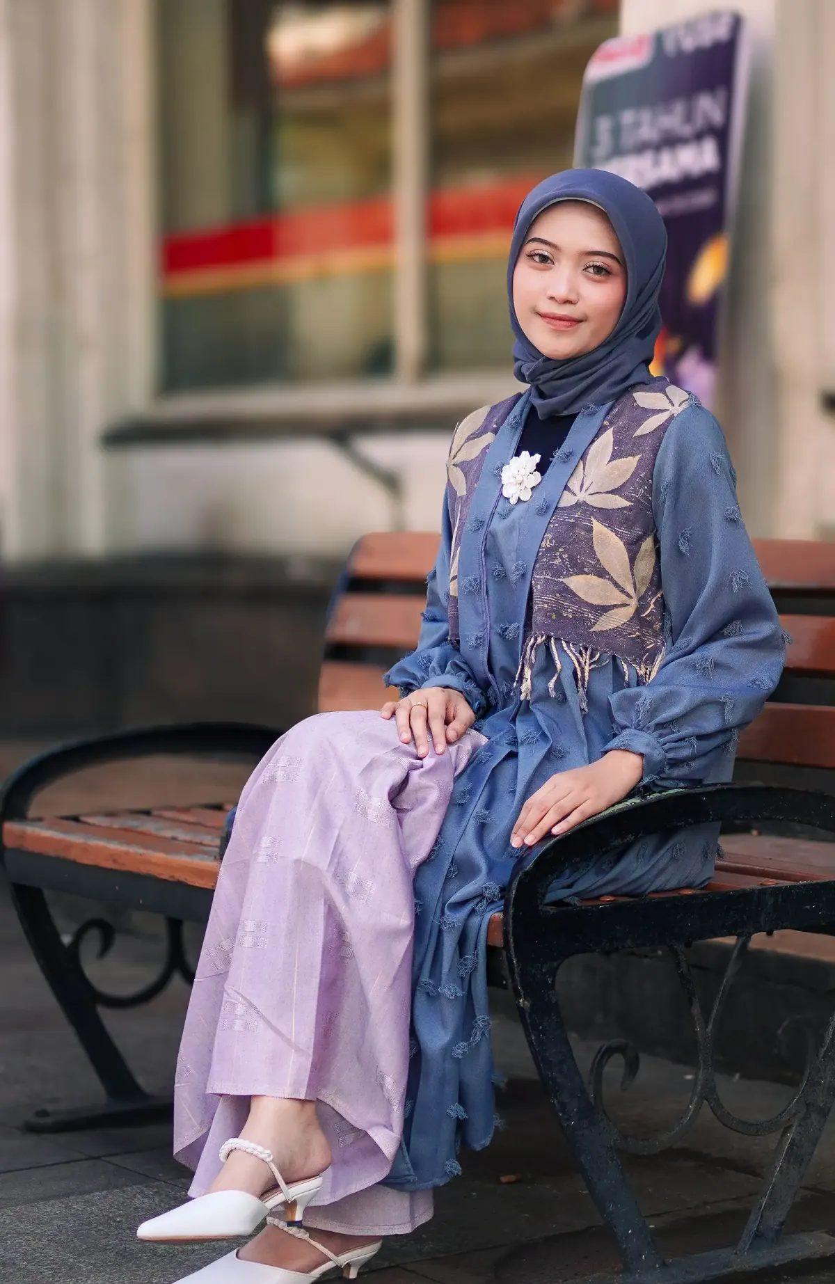 Perempuan berhijab biru keabuan memakai outer biru bertekstur dengan panel motif daun, bros bunga putih, rok panjang ungu muda berkilau, dan sepatu putih bertali mutiara, duduk di bangku taman kota, Bandung.