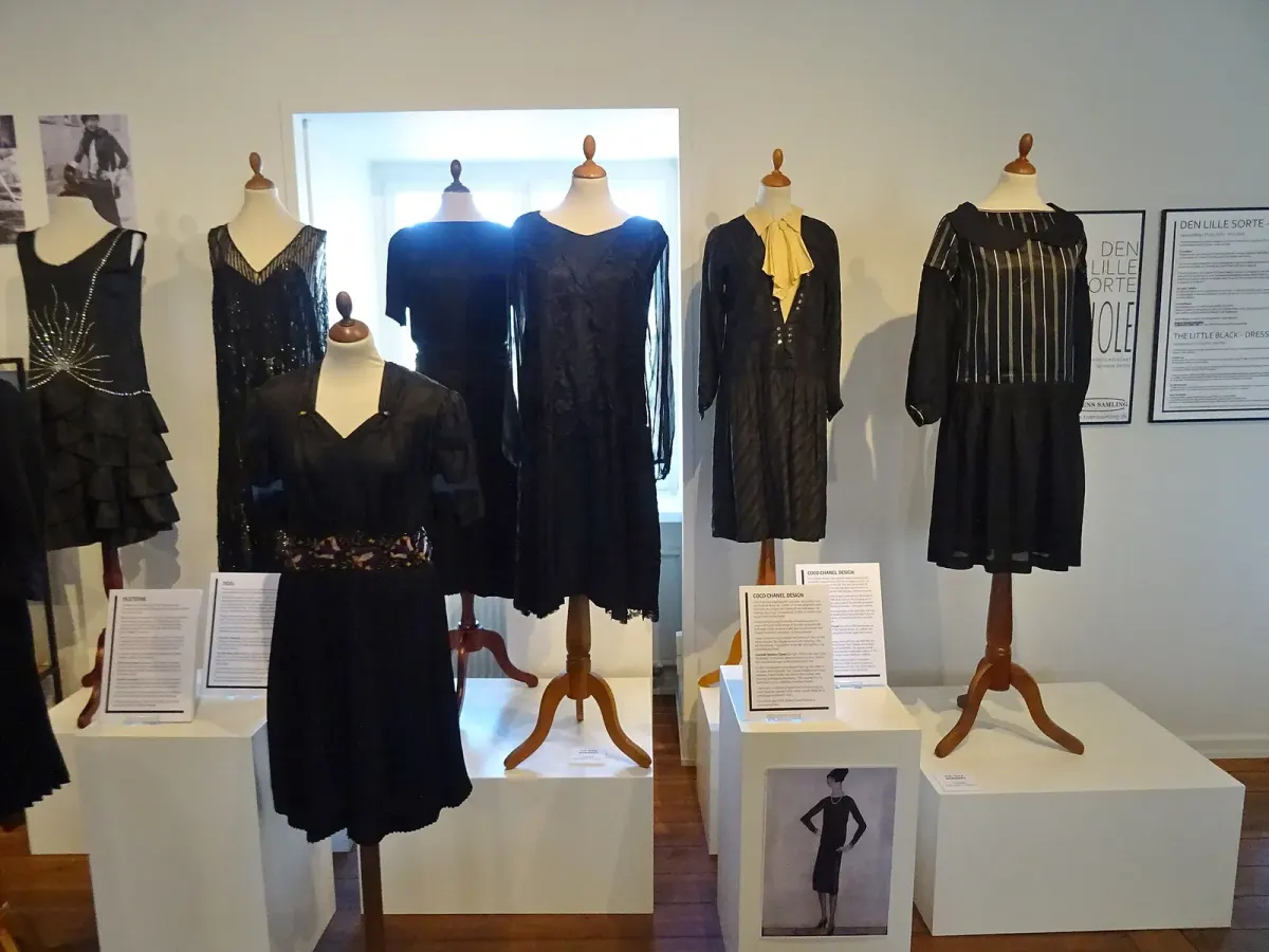Enam dress hitam bergaya 1920-an dipajang di manekin kayu dalam pameran museum, lengkap dengan papan keterangan dan poster pameran di dinding putih.