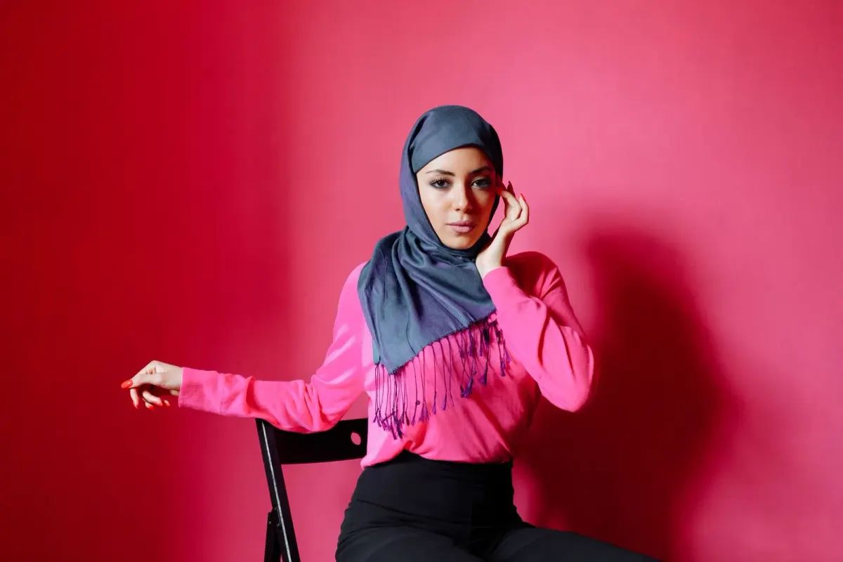 Perempuan duduk di kursi memakai kerudung abu kebiruan dengan rumbai ungu yang melingkari leher dan ujungnya jatuh di depan, atasan merah muda, latar dinding merah.