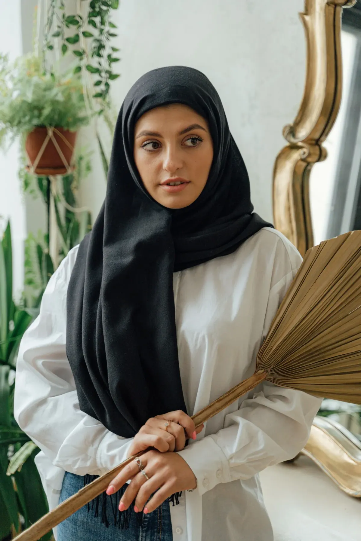 Perempuan memakai kerudung panjang hitam berumbai yang menjuntai menutup dada, kemeja putih longgar, dan celana jeans, memegang daun palem kering.