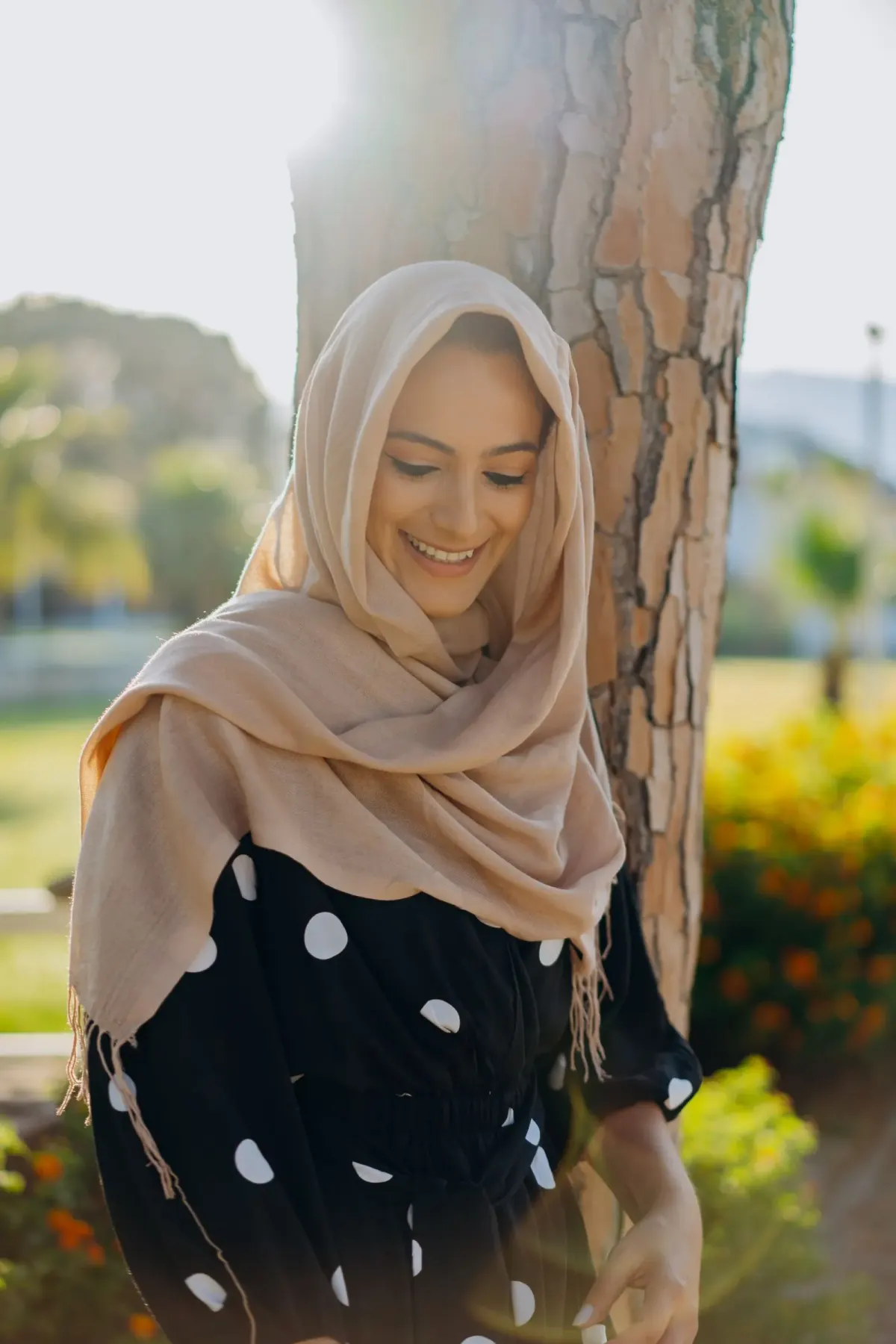 Perempuan tersenyum memakai kerudung panjang krem berbahan tipis bertekstur kusut yang dililitkan longgar ke bahu, dengan baju hitam bermotif polkadot putih.