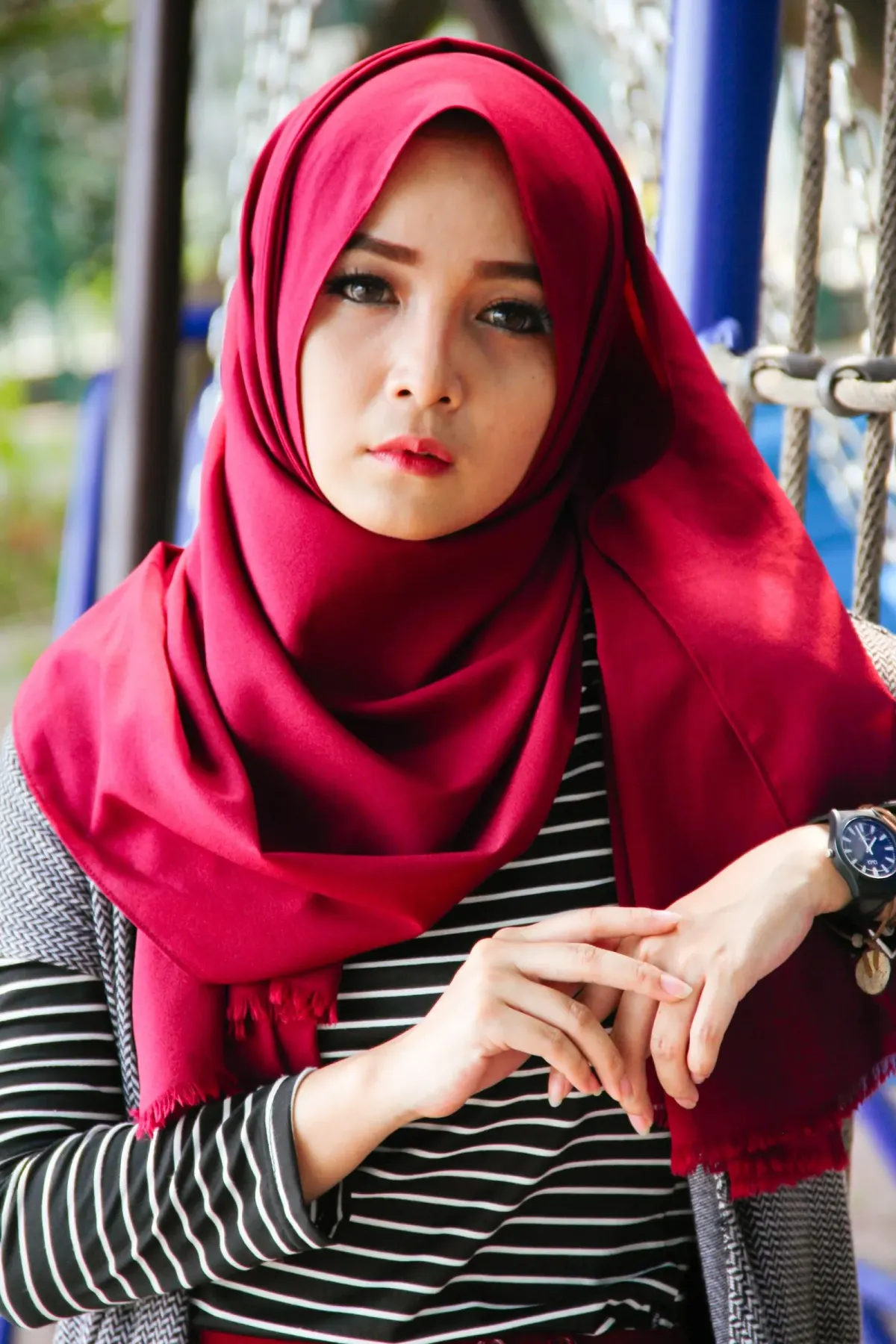 Perempuan memakai kerudung panjang merah berumbai yang satu sisinya dililitkan ke bahu, dipadukan kaus bergaris hitam putih, kardigan abu, dan jam tangan.