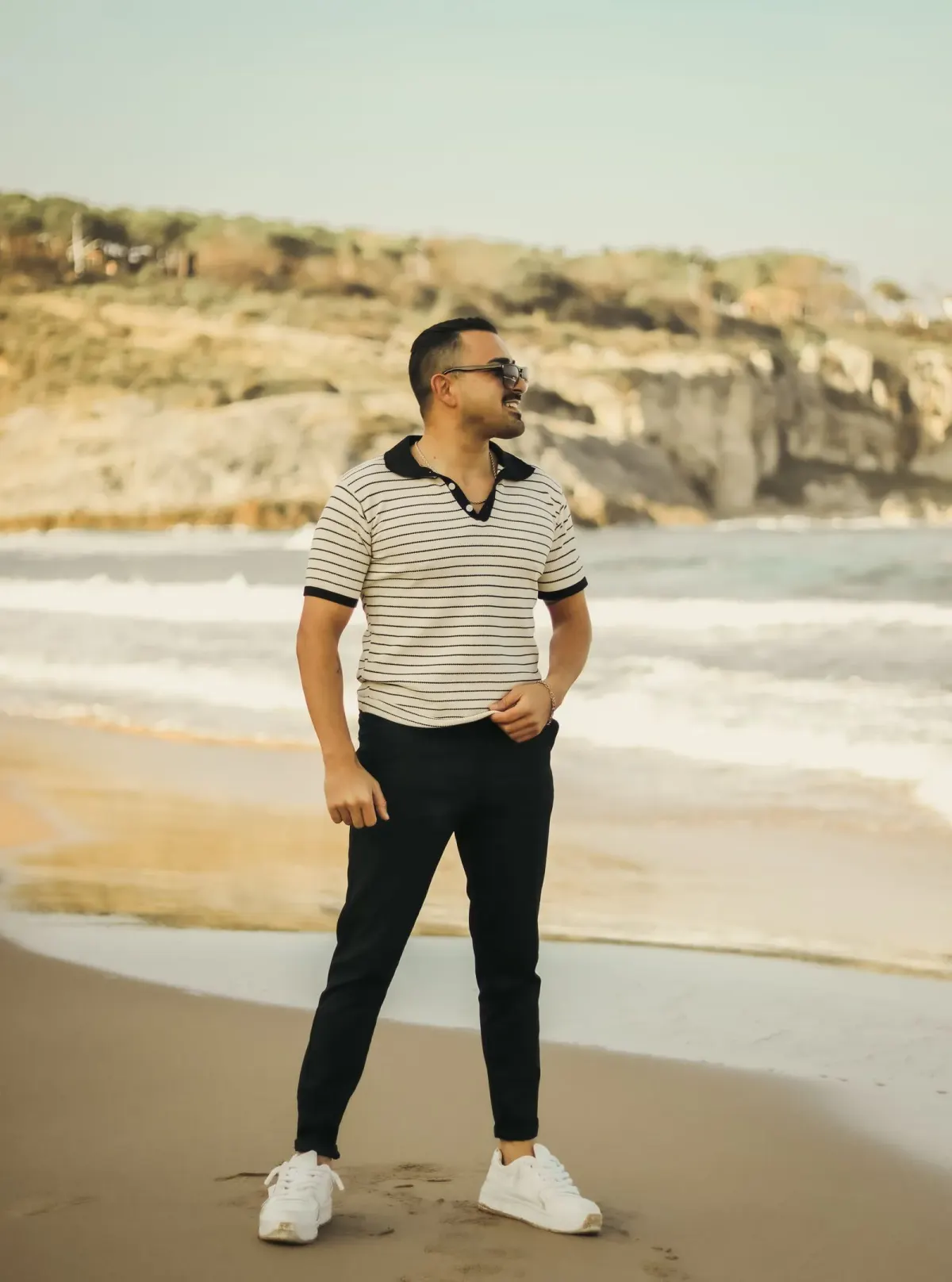 Pria berkacamata hitam memakai polo rajut krem bergaris dengan kerah hitam, celana hitam slim, dan sneakers putih, berdiri di pantai dengan tebing di belakang.