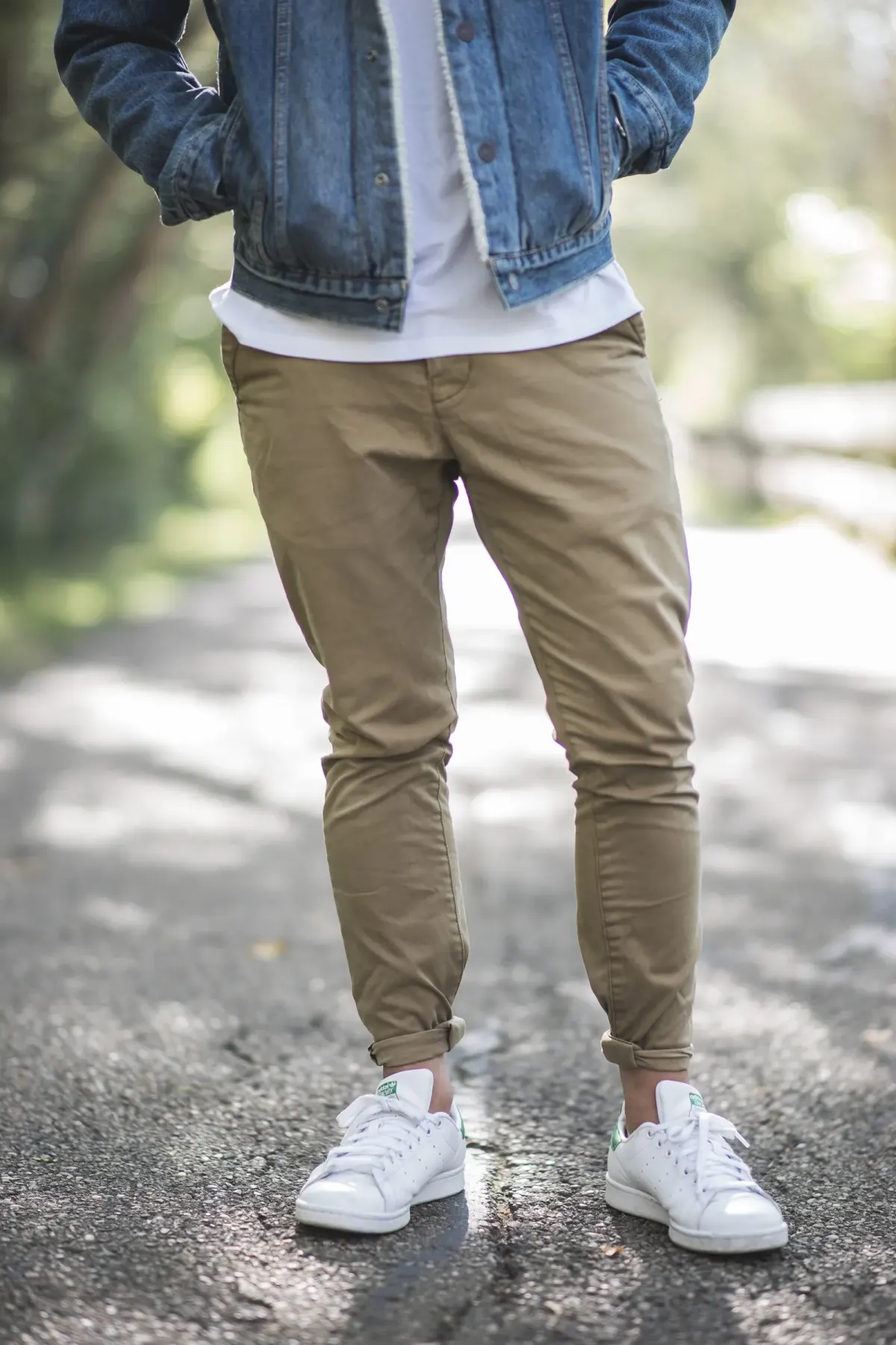 Pria memakai jaket denim biru berlapis bulu putih di bagian dalam, kaus putih polos, celana chino khaki yang ujungnya digulung, dan sneakers kulit putih, berdiri di jalan aspal. Wajah tidak terlihat.