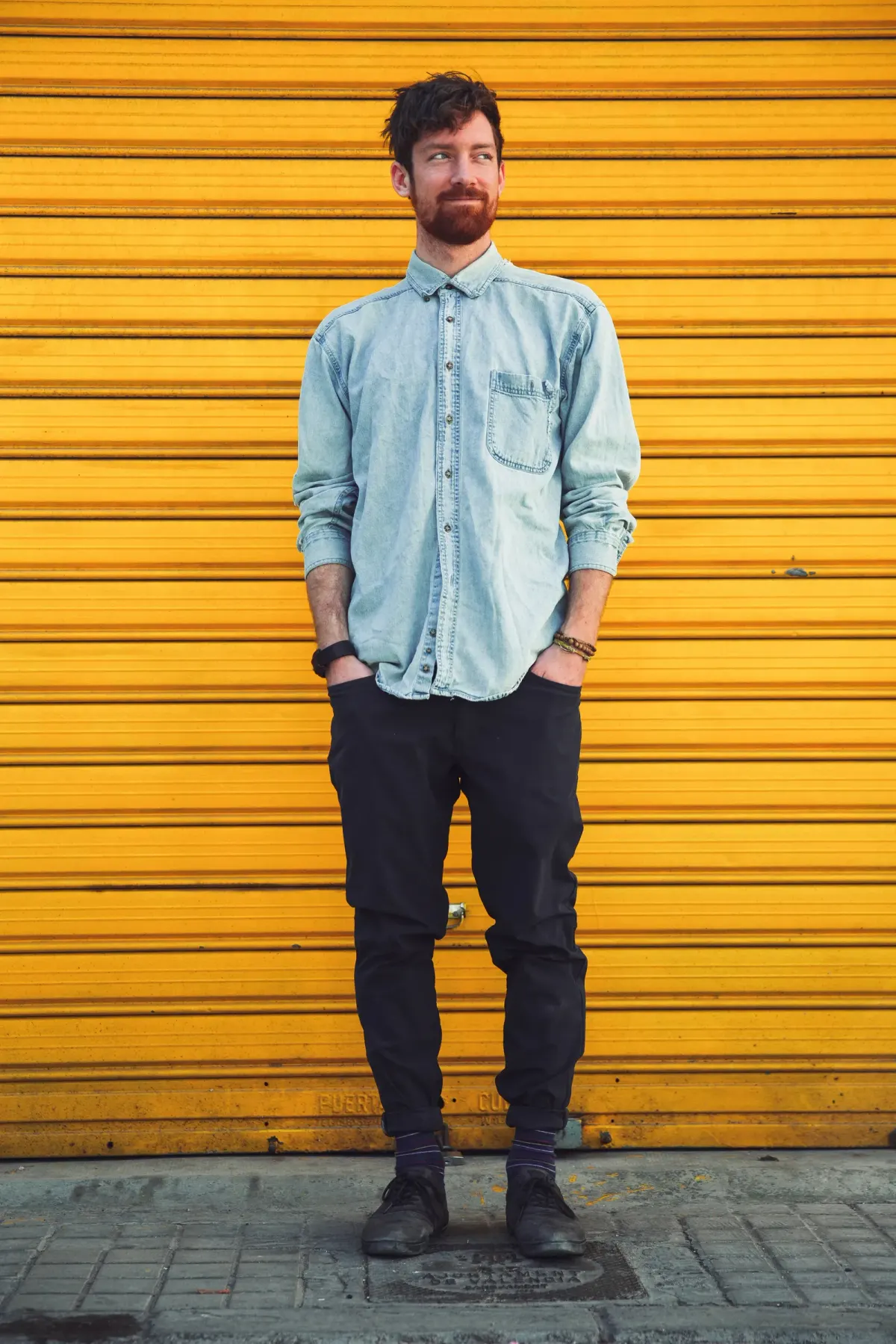Pria berjanggut memakai kemeja denim biru muda berkantong dada dengan lengan digulung, celana hitam yang mengecil di pergelangan kaki, dan sepatu hitam, berdiri di depan rolling door kuning.