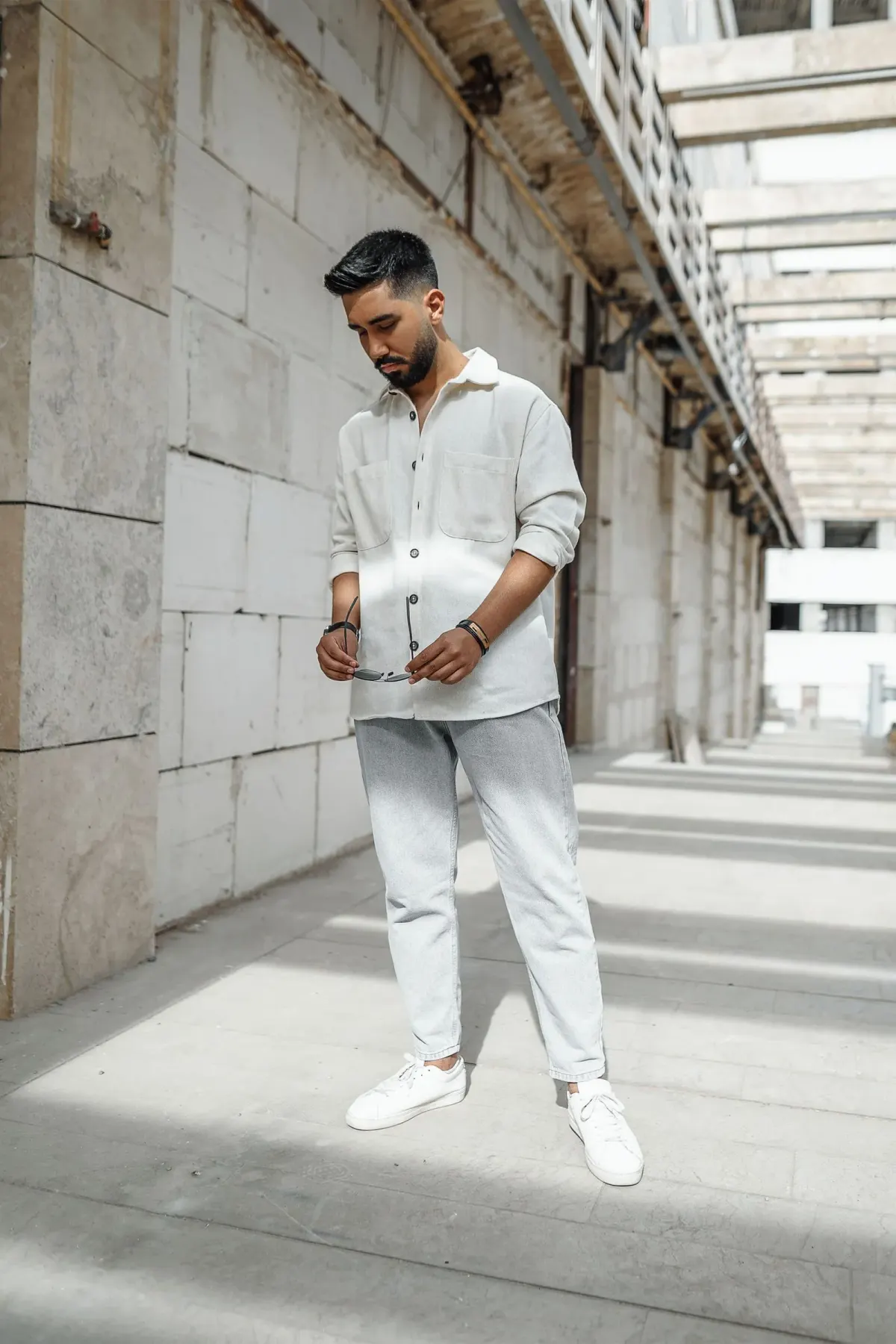 Pria berdiri di lorong beton memakai kemeja putih gading berlengan digulung dengan dua saku dada, celana abu-abu muda, sneakers low-top putih, dan gelang hitam, sambil memegang kacamata hitam.