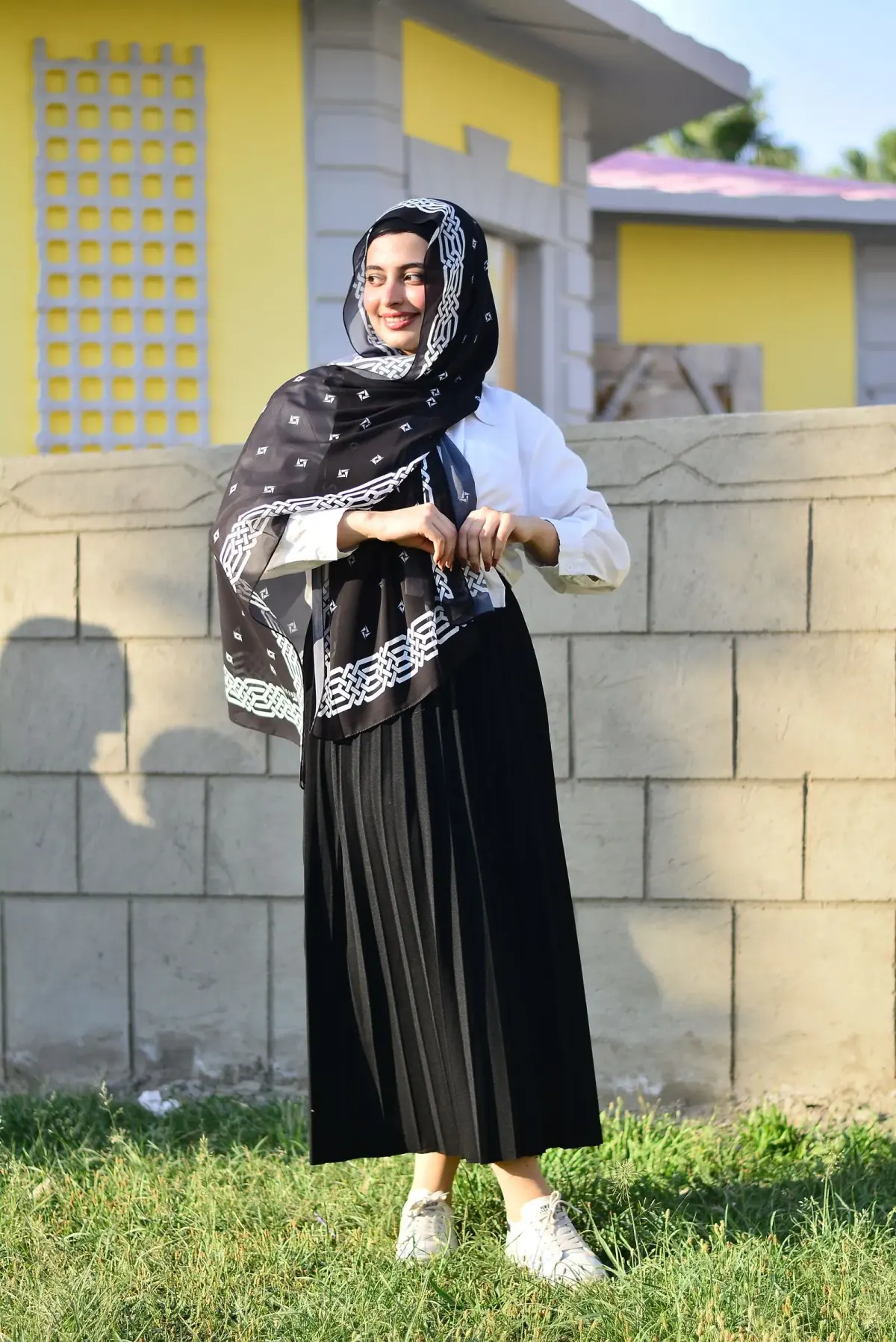 Perempuan berhijab memakai selendang hitam bermotif putih, kemeja putih lengan panjang, rok plisket hitam panjang, dan sneakers putih, berdiri di rumput depan tembok batako.